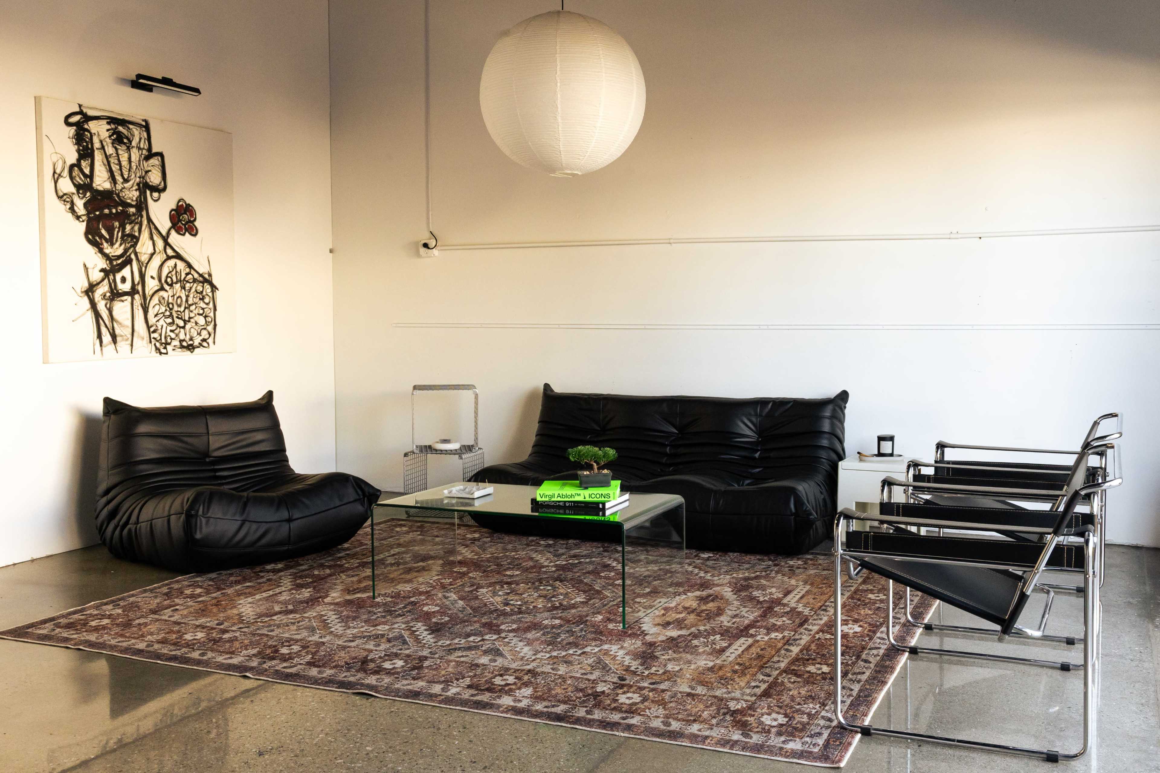 The image shows a modern living space featuring black leather furniture, a glass coffee table, and a patterned area rug under soft lighting.