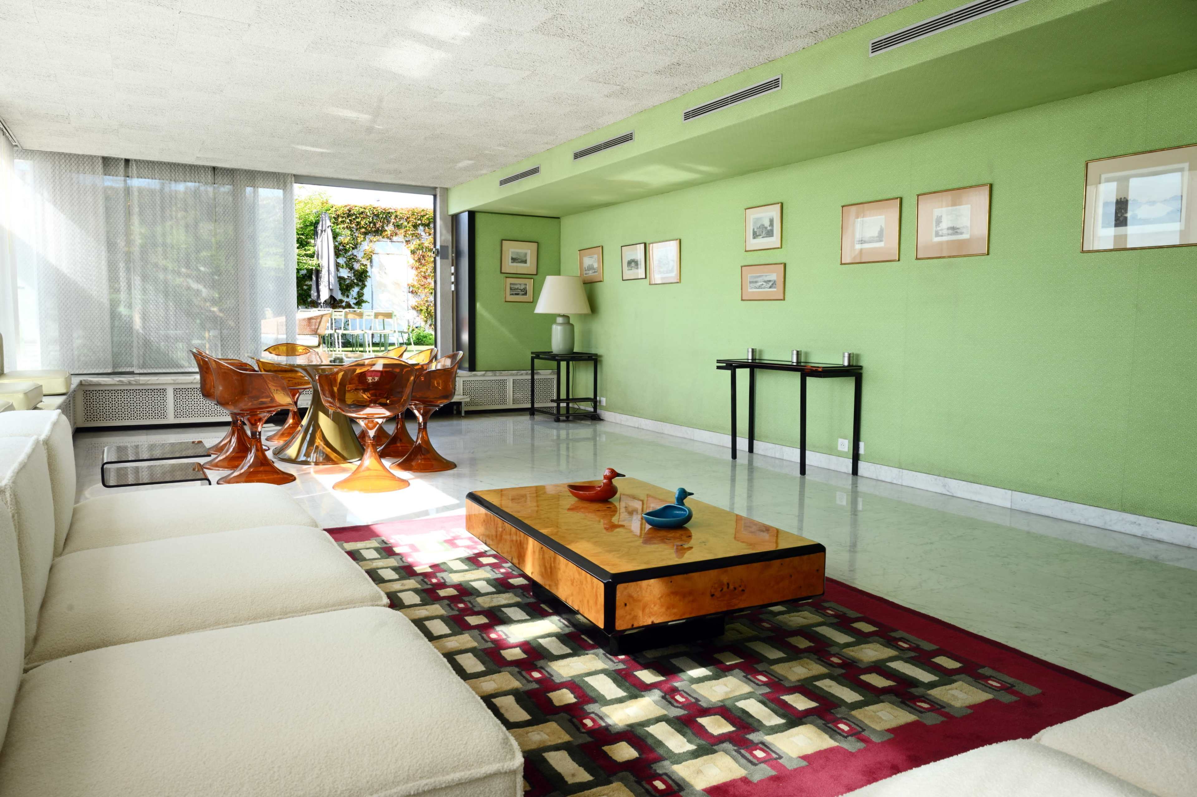 The image shows a modern living room featuring a green wall, a white sofa, a wooden coffee table, and a dining area with transparent chairs and a round table.