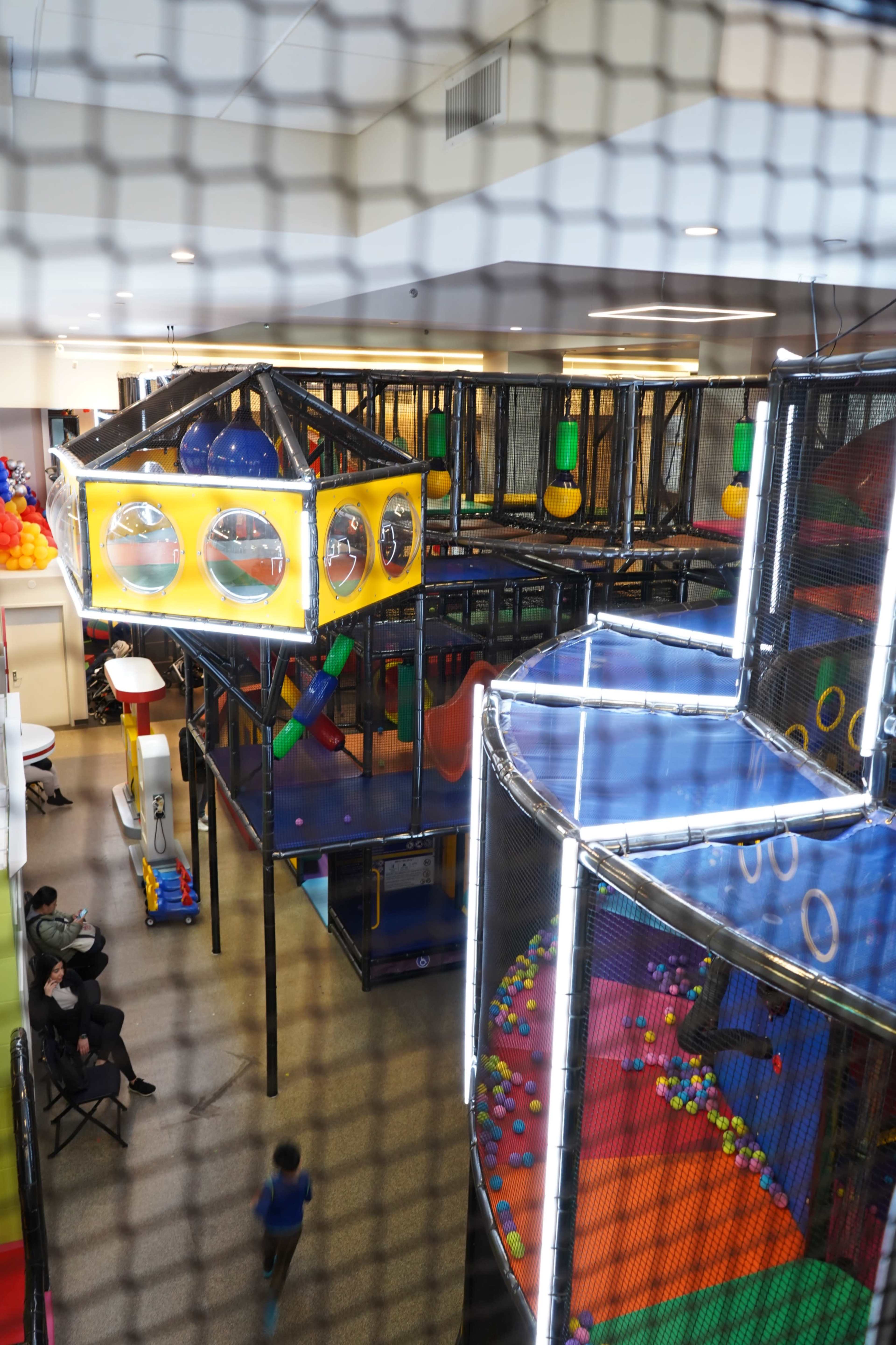 The image shows a colorful indoor play area with various climbing structures, slides, and ball pits enclosed by safety netting.