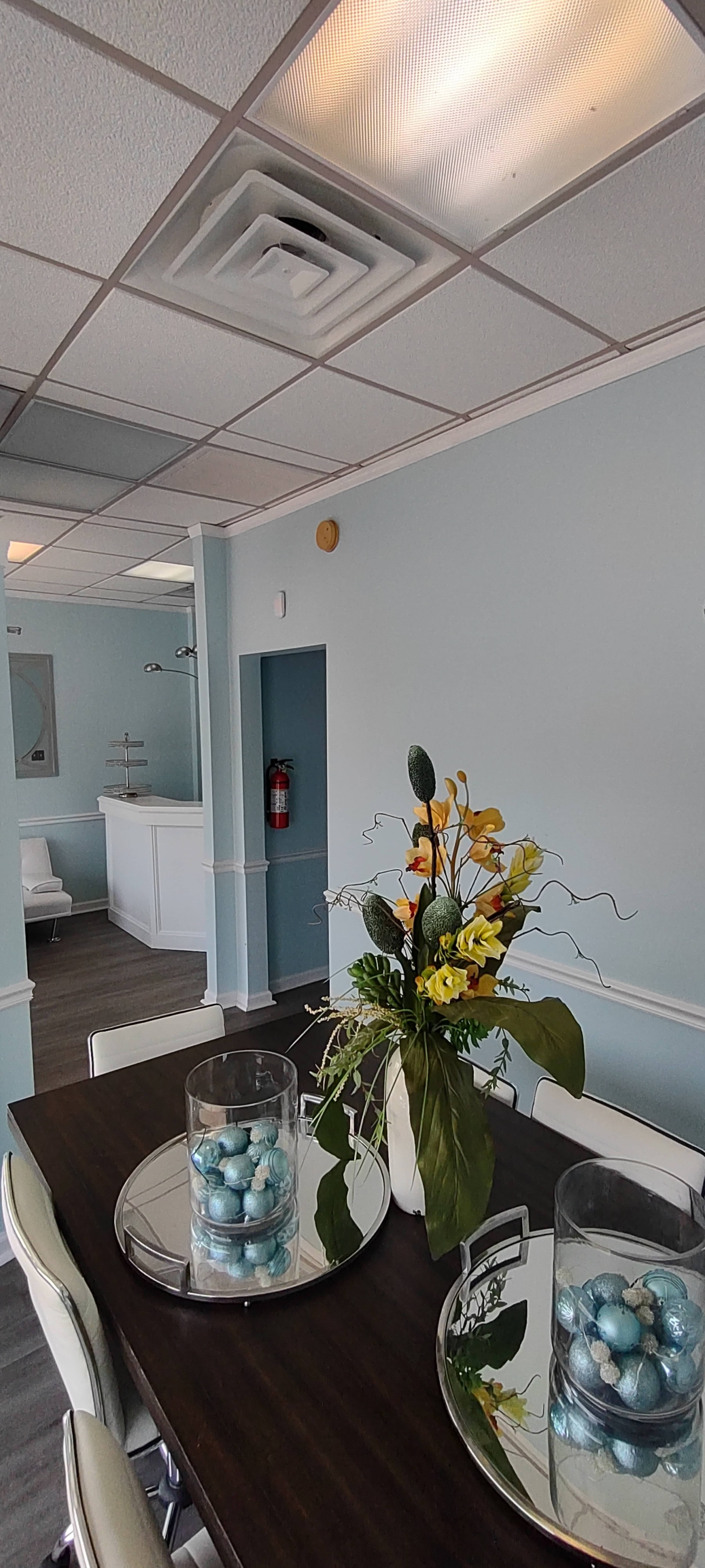 A modern waiting area features a dark wooden table with a floral centerpiece, light blue walls, and a reception counter in the background.