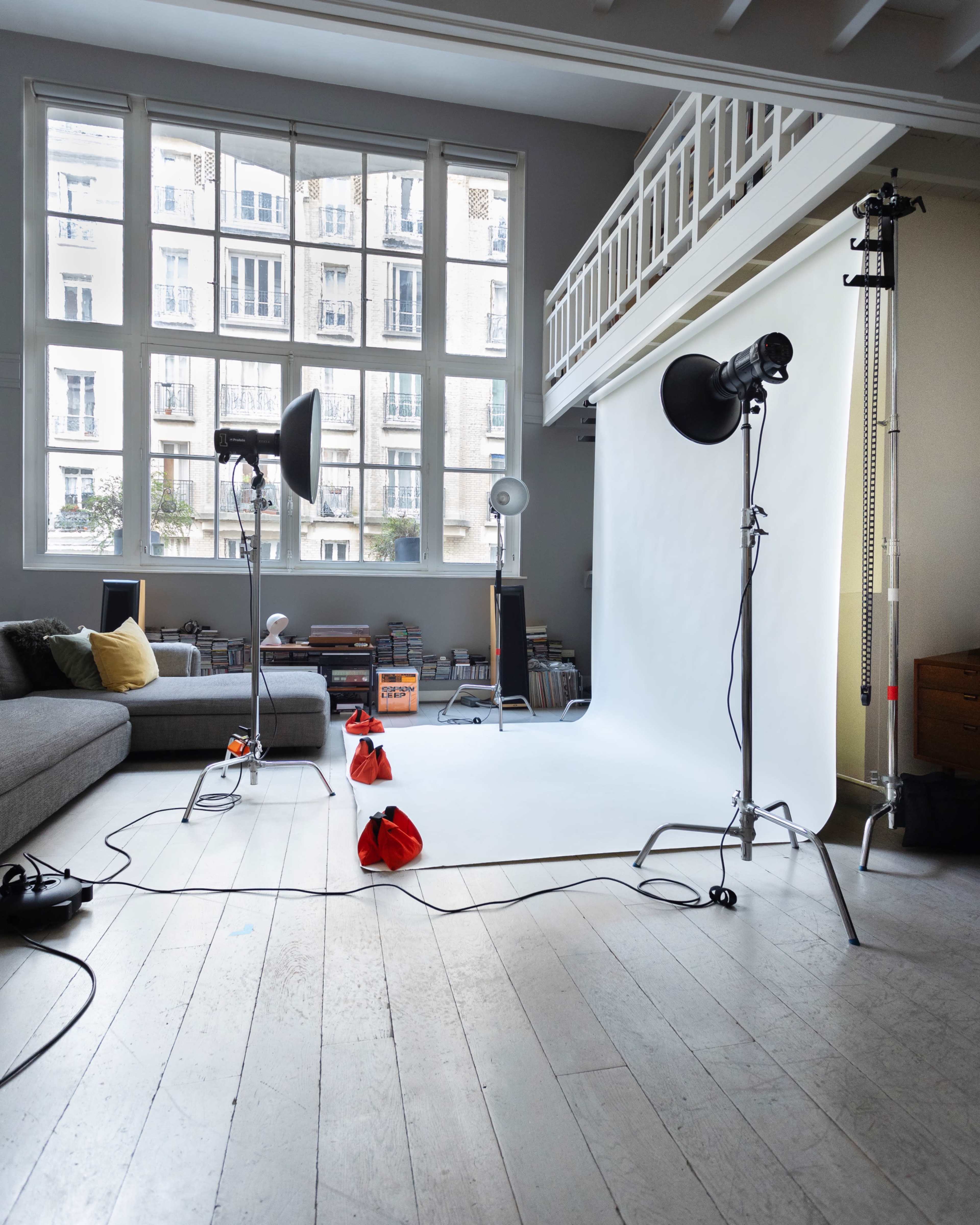 A photography studio setup features a white backdrop, lighting equipment, and props arranged on a wooden floor in a room with large windows.