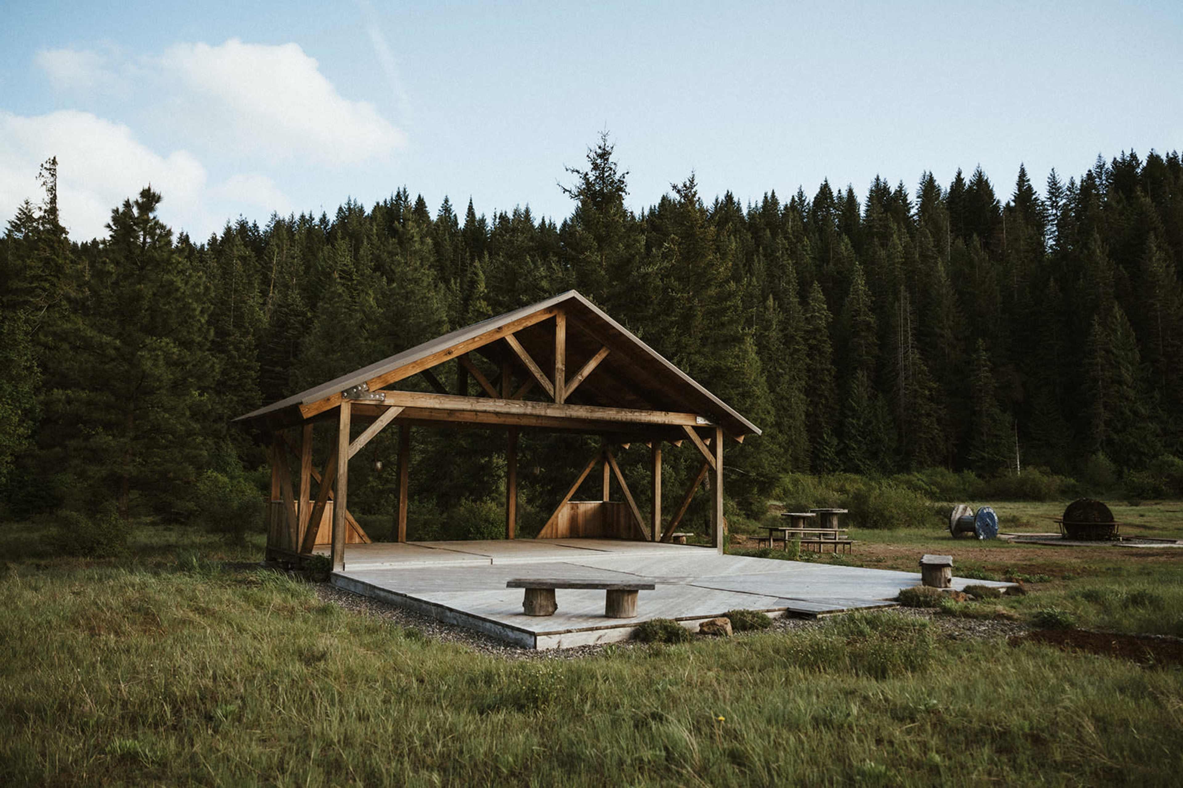 A wooden pavilion stands in a grassy area surrounded by tall trees in a forested setting.