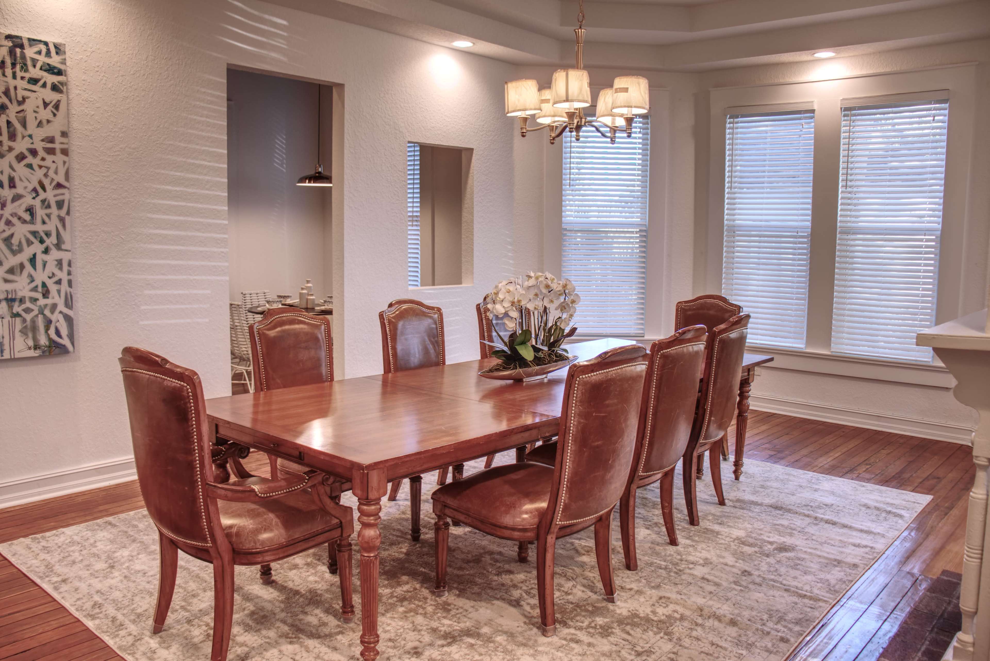 A dining room features a long wooden table surrounded by eight leather chairs, with a chandelier overhead and large windows allowing natural light to enter.