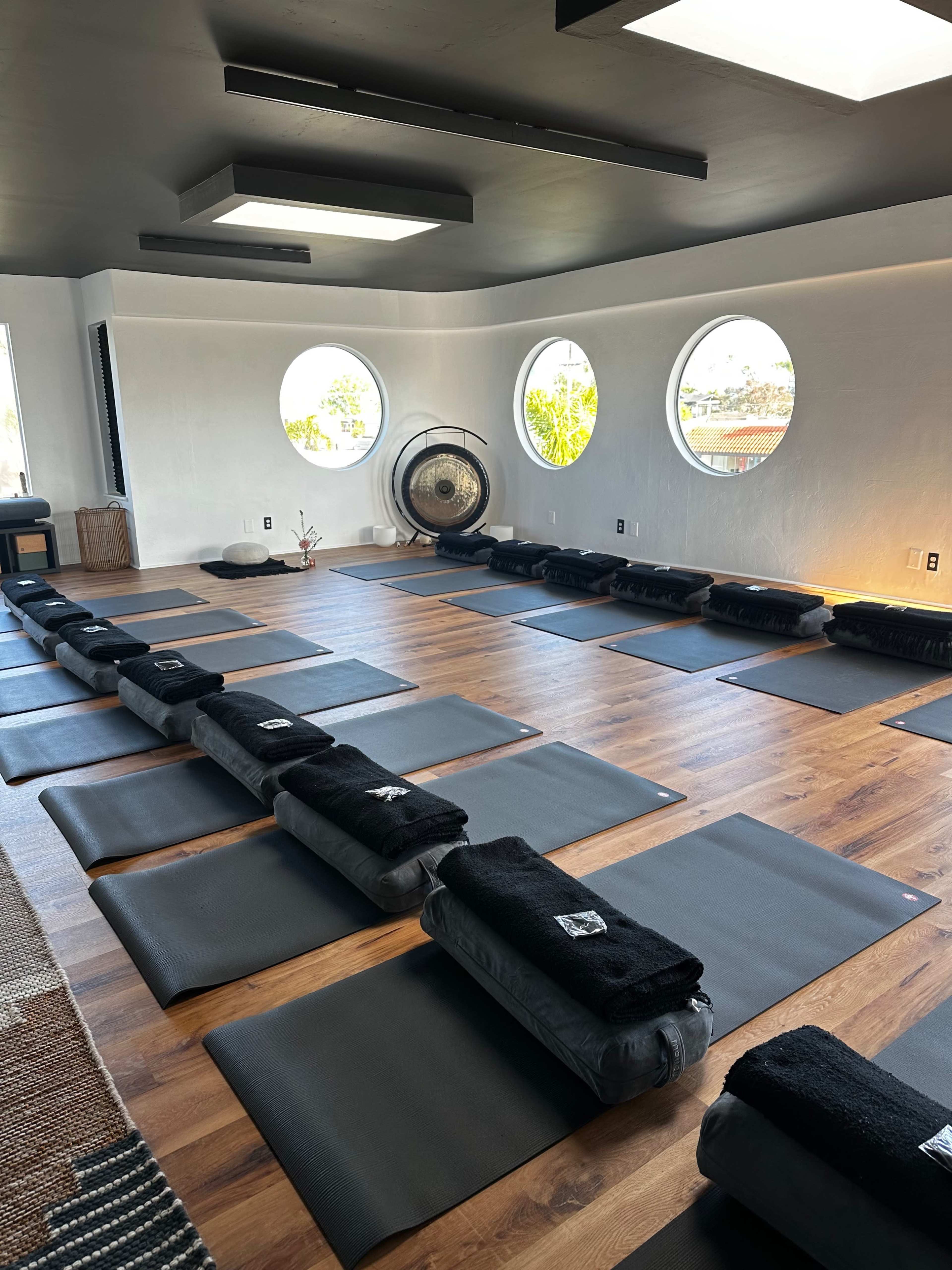 The image shows a spacious yoga studio with neatly arranged mats and rolled towels, complemented by large circular windows that allow natural light to enter.
