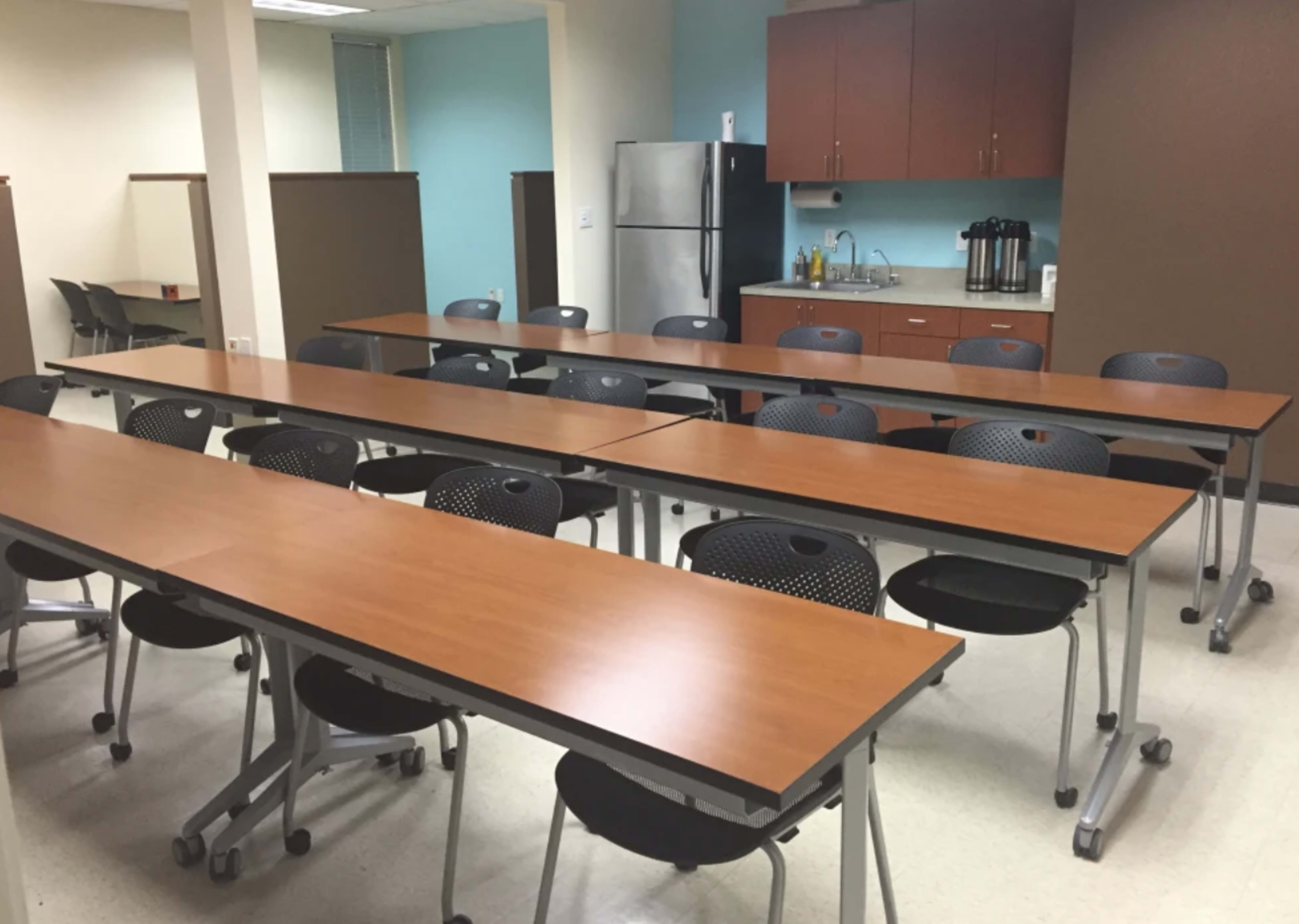 A meeting room features several tables arranged in rows, accompanied by rolling chairs and a kitchenette area at the back.