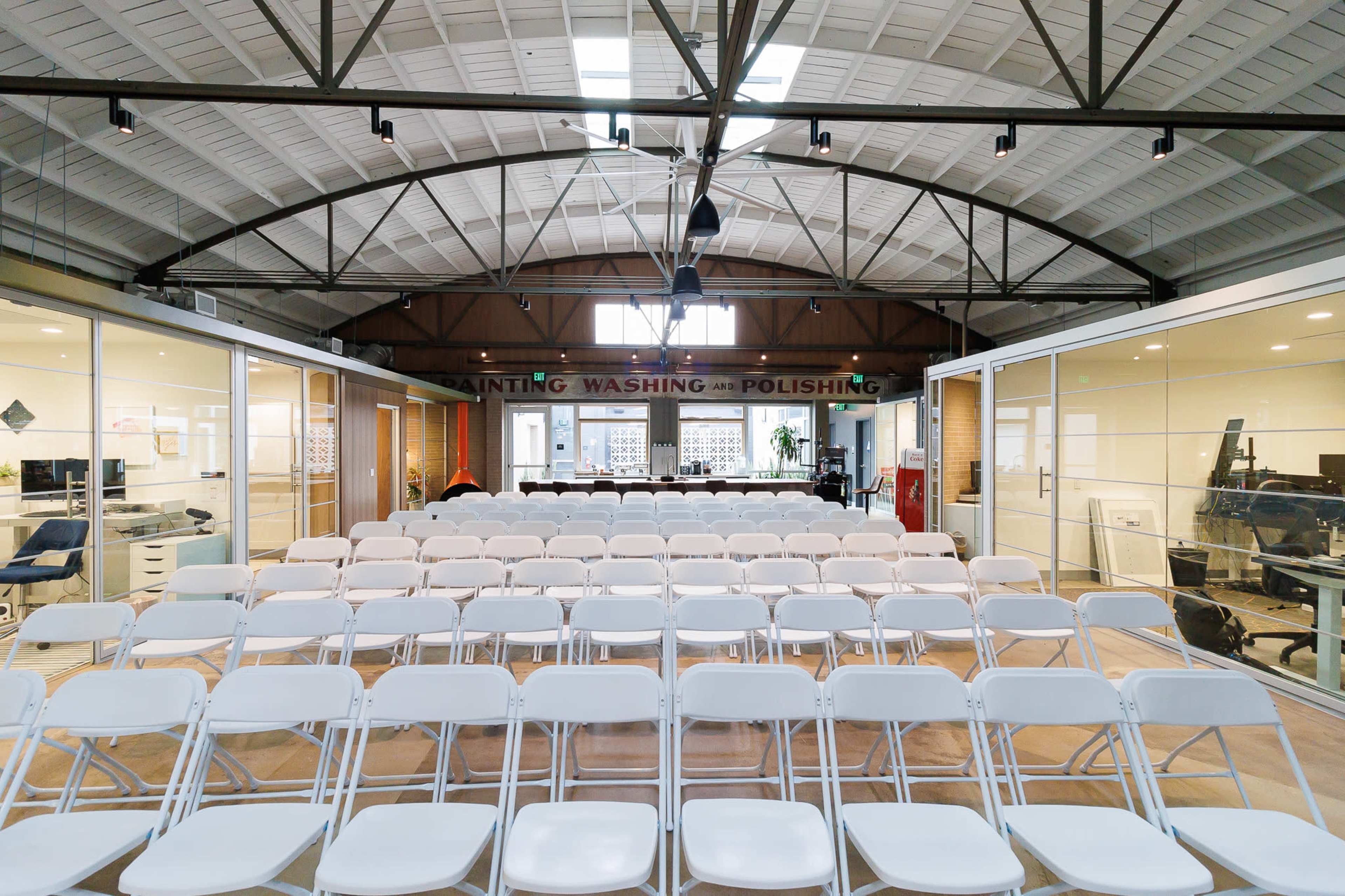 A spacious room with rows of white chairs facing a table in a well-lit area featuring large windows and high ceilings.