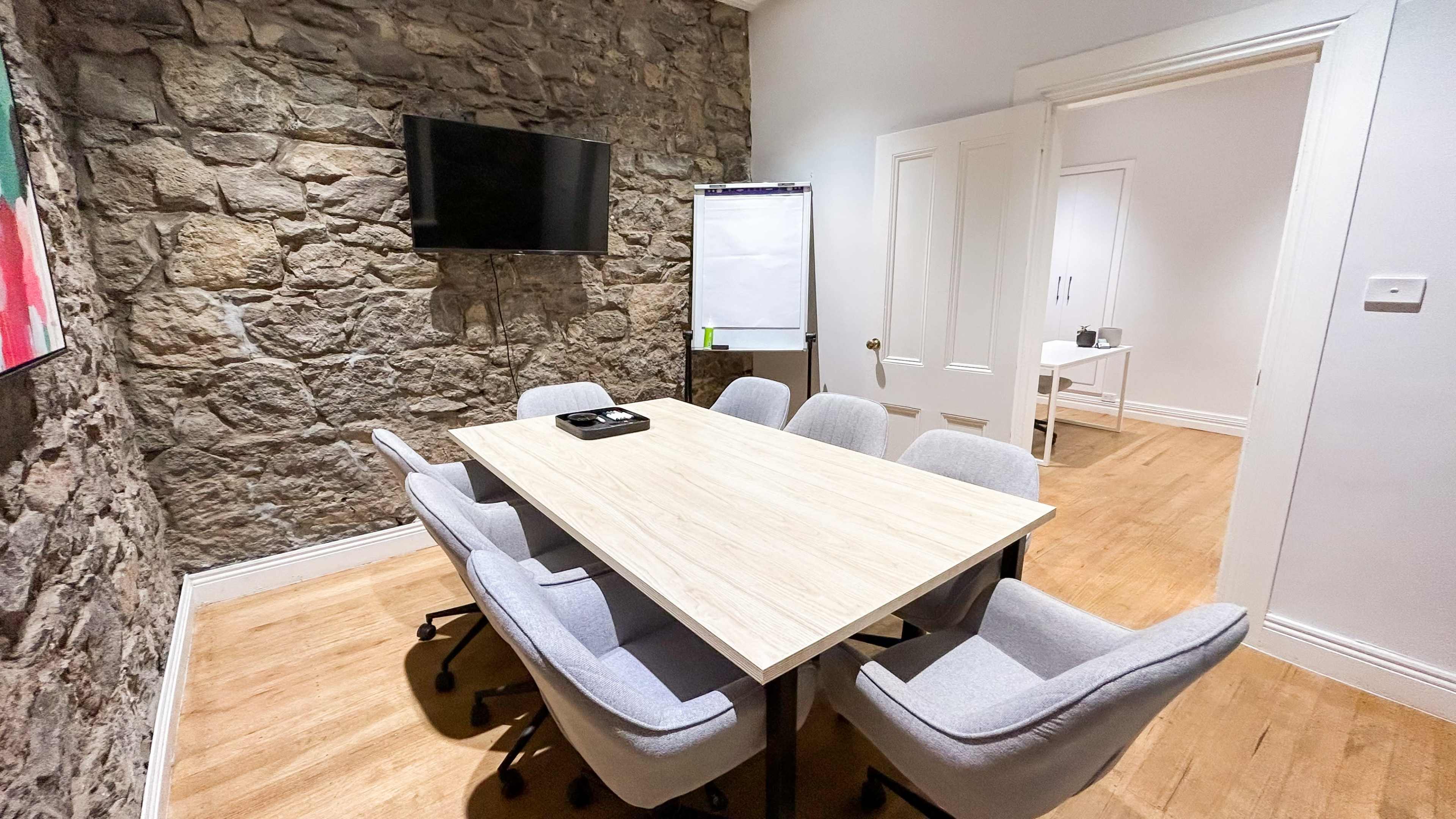 A conference room features a wooden table surrounded by gray chairs, a wall of exposed stone, a television mounted on the wall, and a whiteboard nearby.