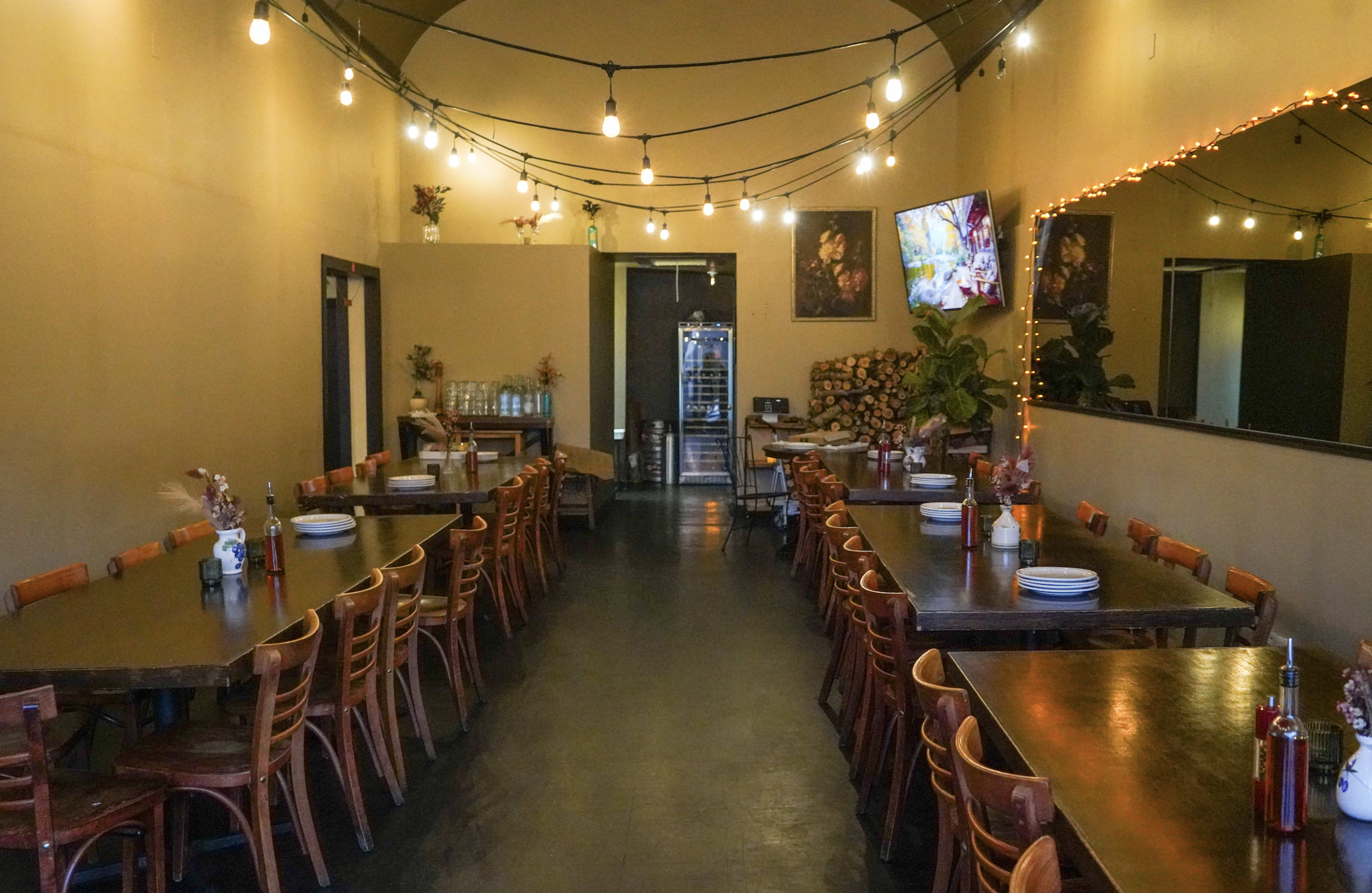 The image shows a spacious restaurant interior with several wooden dining tables arranged in rows, string lights hanging from the ceiling, and a counter visible in the background.