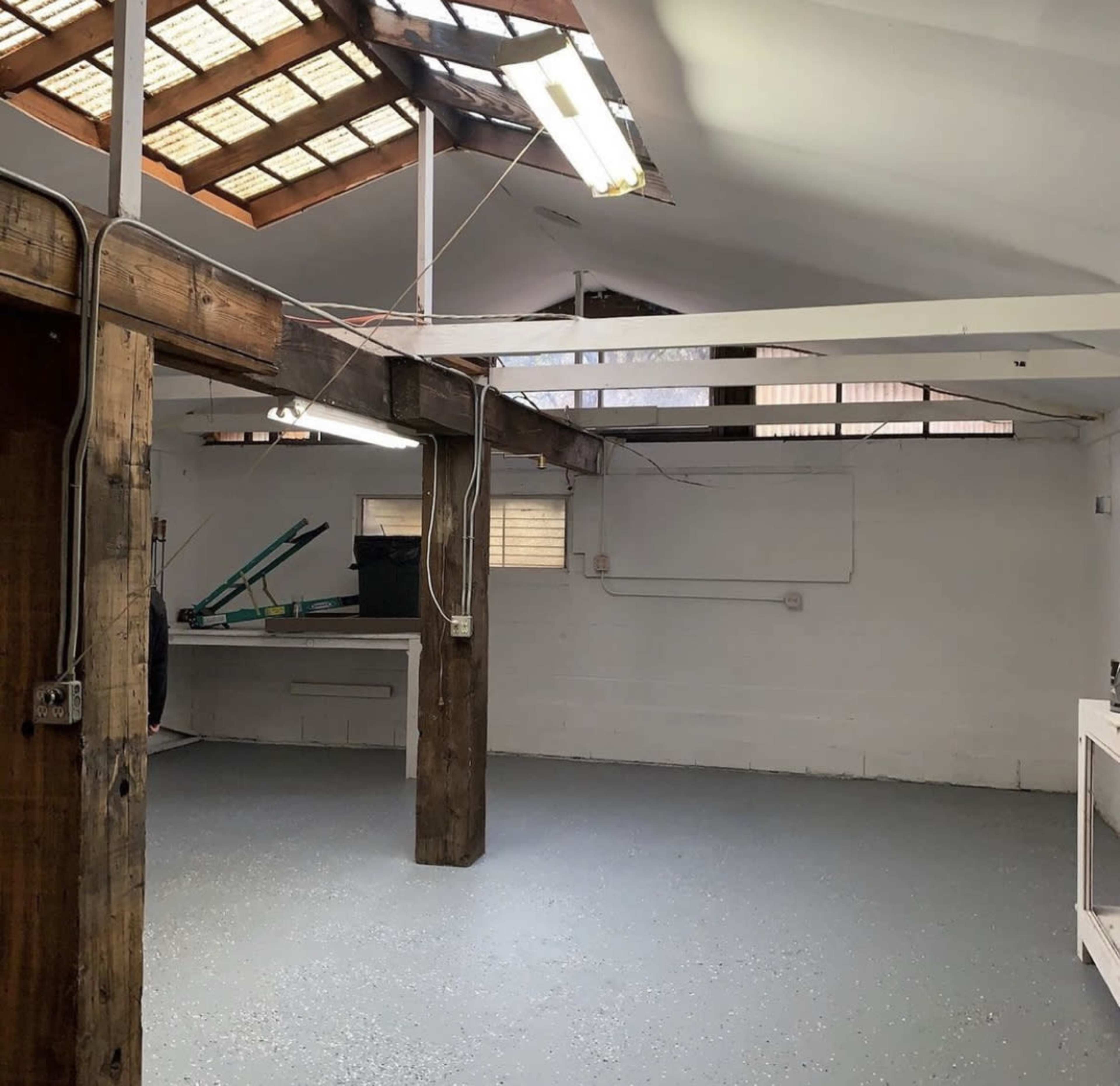 The image shows an empty room with wooden beams, a gray floor, and overhead lighting, featuring a work area with some equipment in the background.