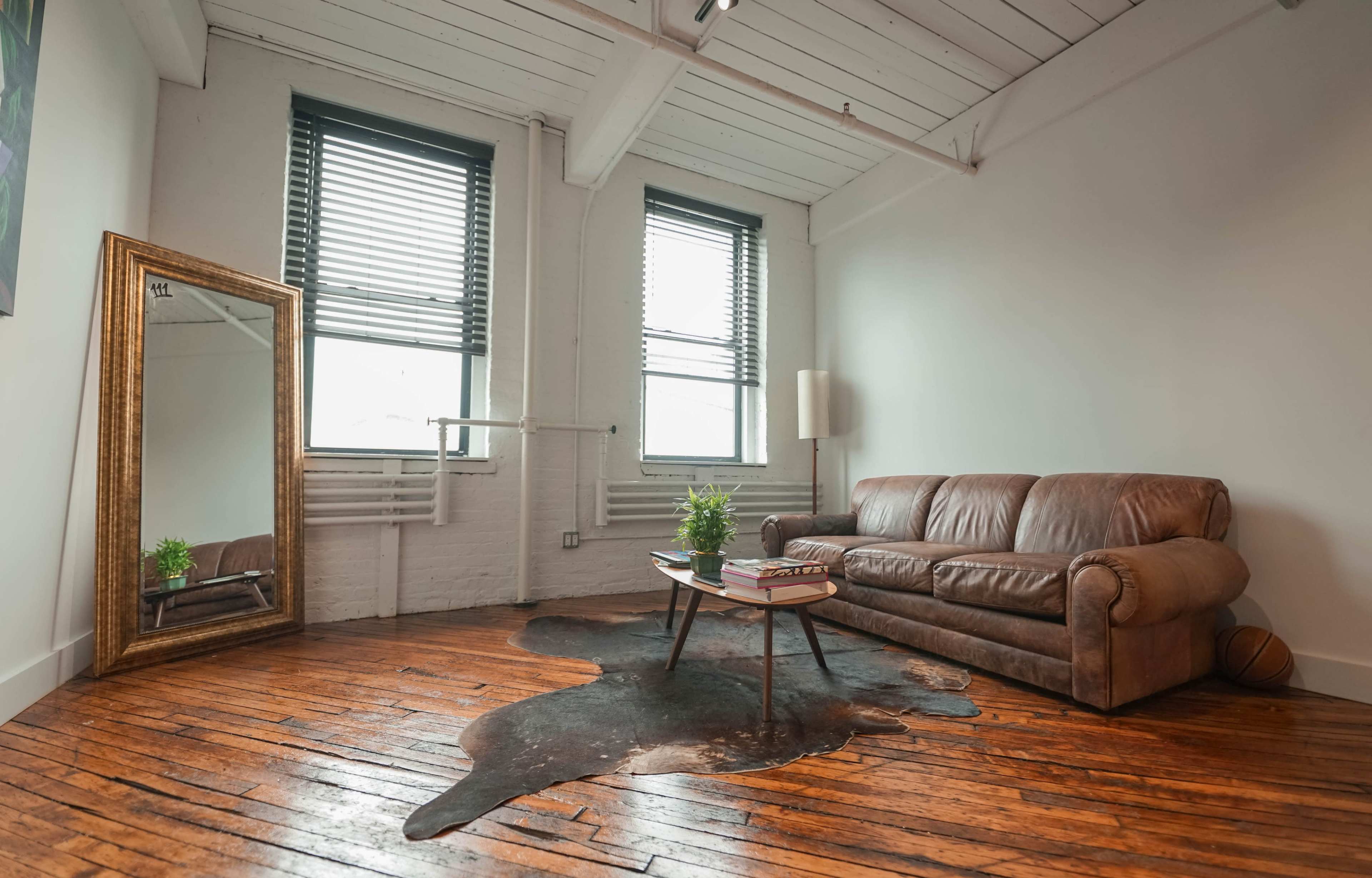A minimalist living room featuring a brown leather sofa, a coffee table with books, a cowhide rug on the wooden floor, and a large framed mirror against the wall.