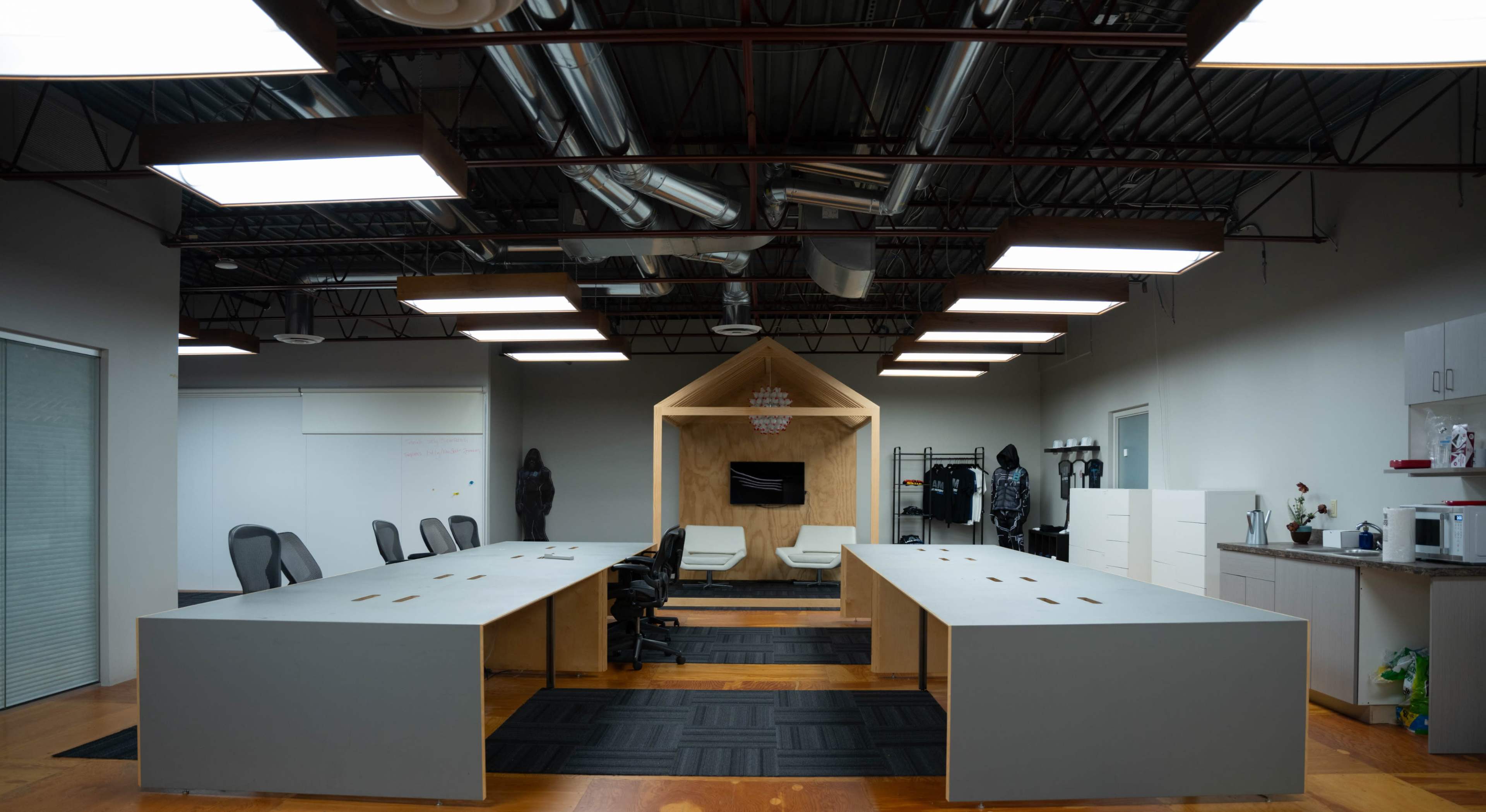 The image shows a modern office space with two large, gray desks facing a small wooden structure that contains seating and a television.