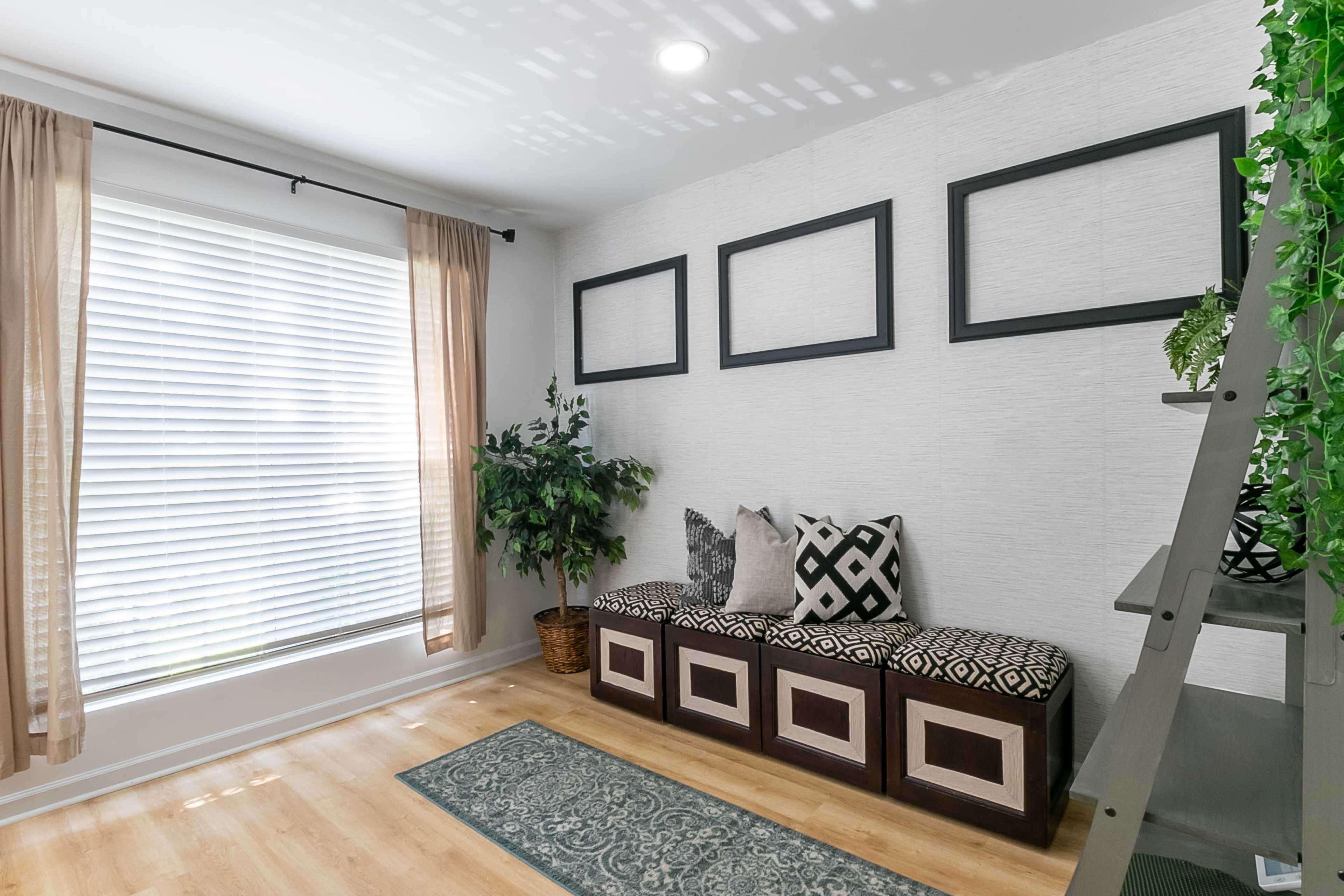 A small, well-lit corner of a room with a window dressed in curtains, a seating area with patterned cushions, and a plant beside a decorative shelving unit.