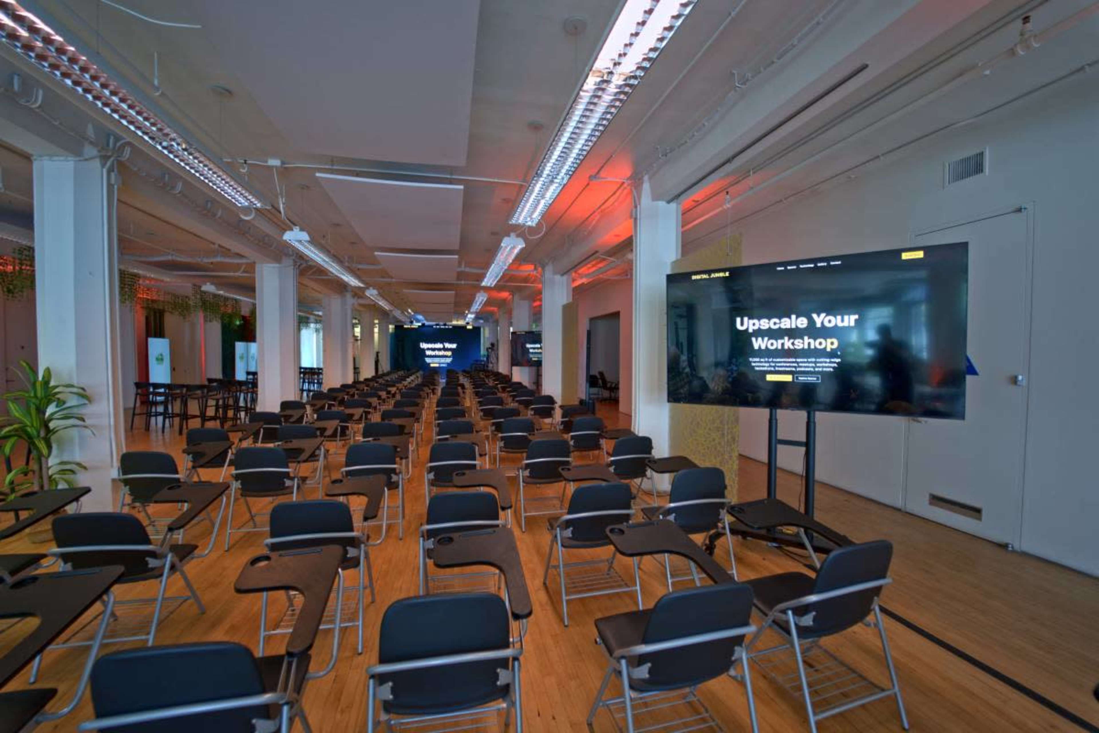 The image shows a spacious room arranged with rows of empty chairs facing large screens displaying information about a workshop.