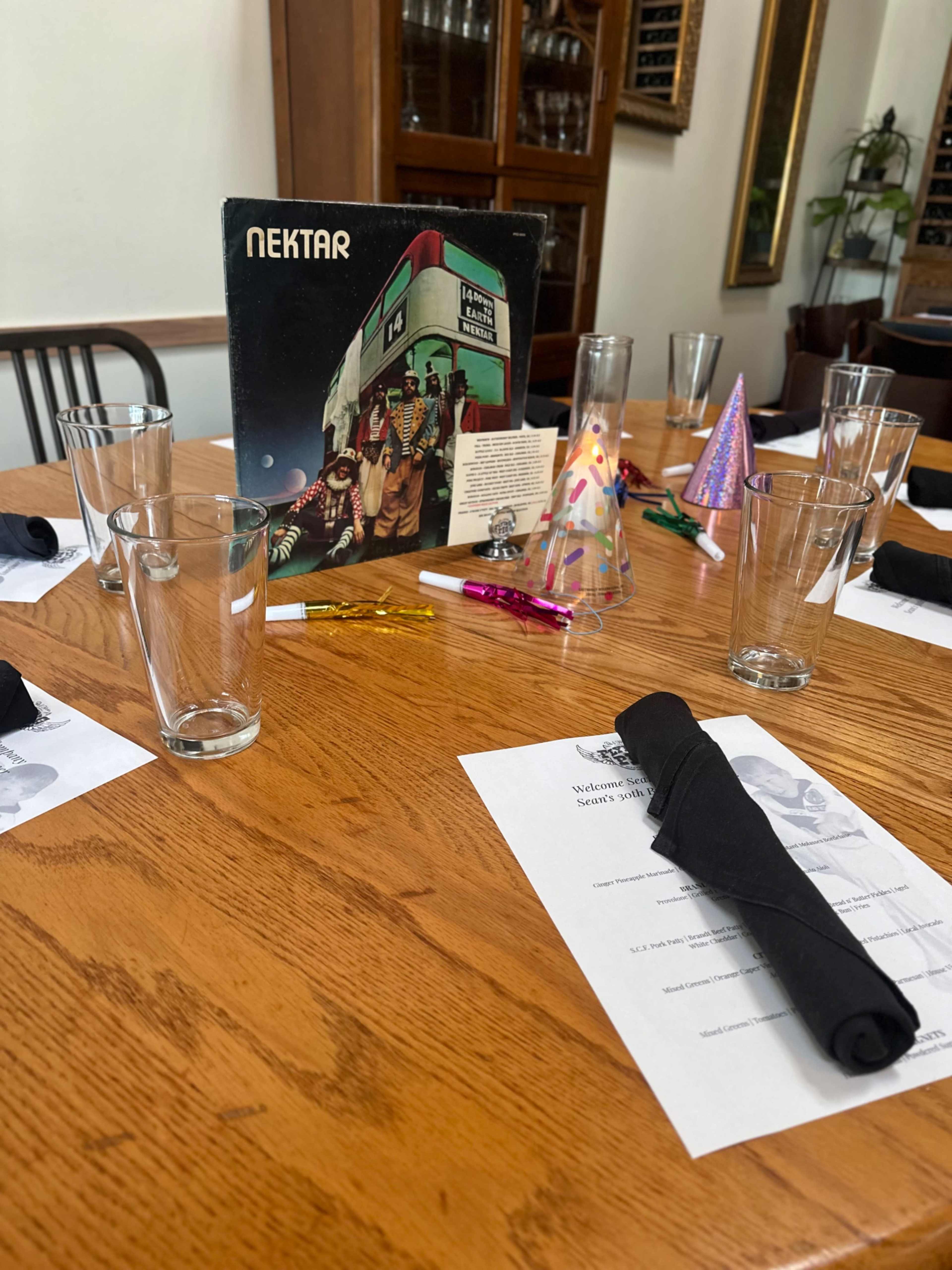 A wooden dining table is set with glasses, napkins, party hats, and a record album at the center.