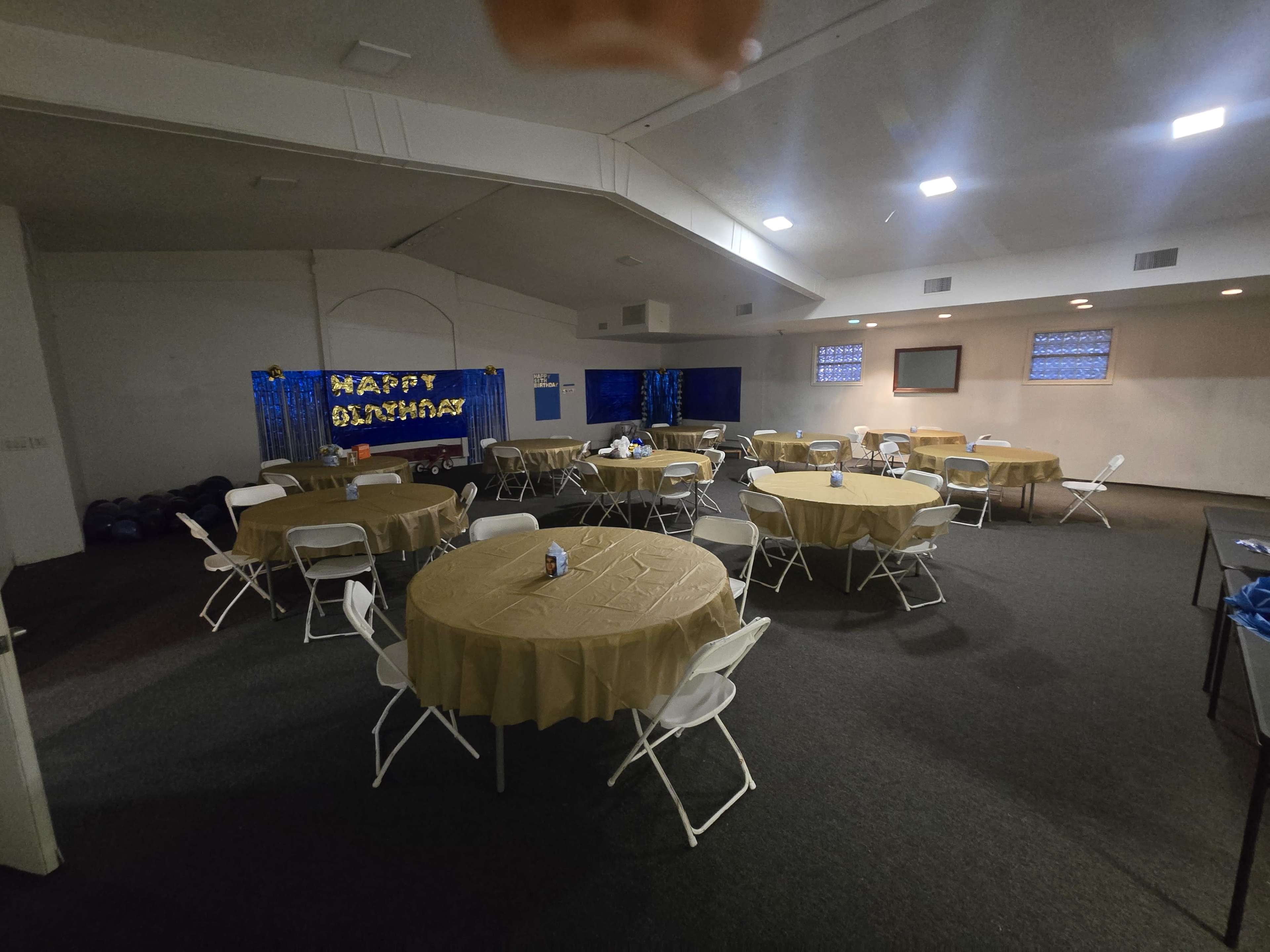 The image shows a room set up for an event, featuring several round tables covered with beige tablecloths and decorated with a "Happy Birthday" banner in the background.