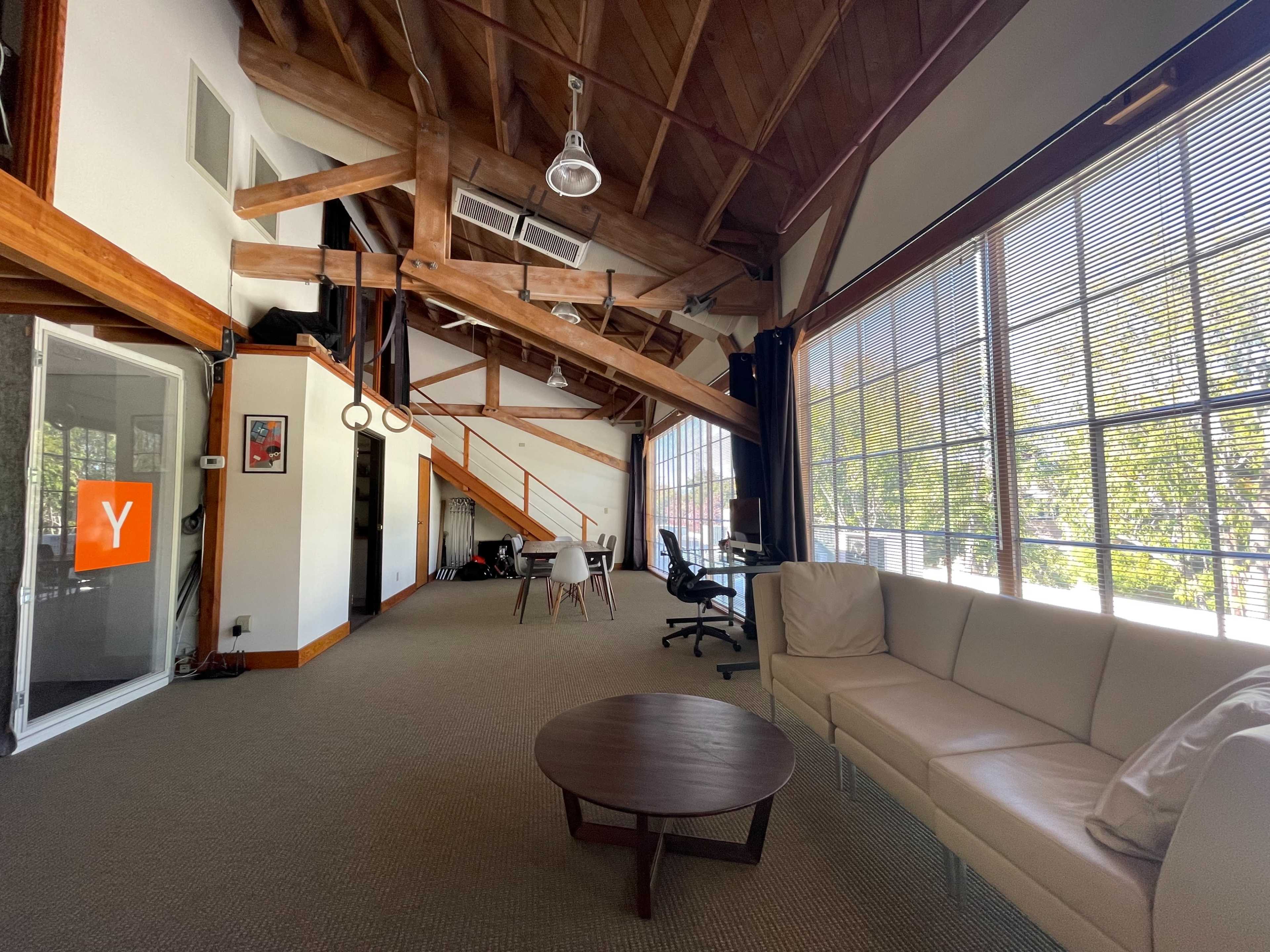 A spacious room with exposed wooden beams, large windows, minimal furniture including a couch and a coffee table, and office workstations in the background.