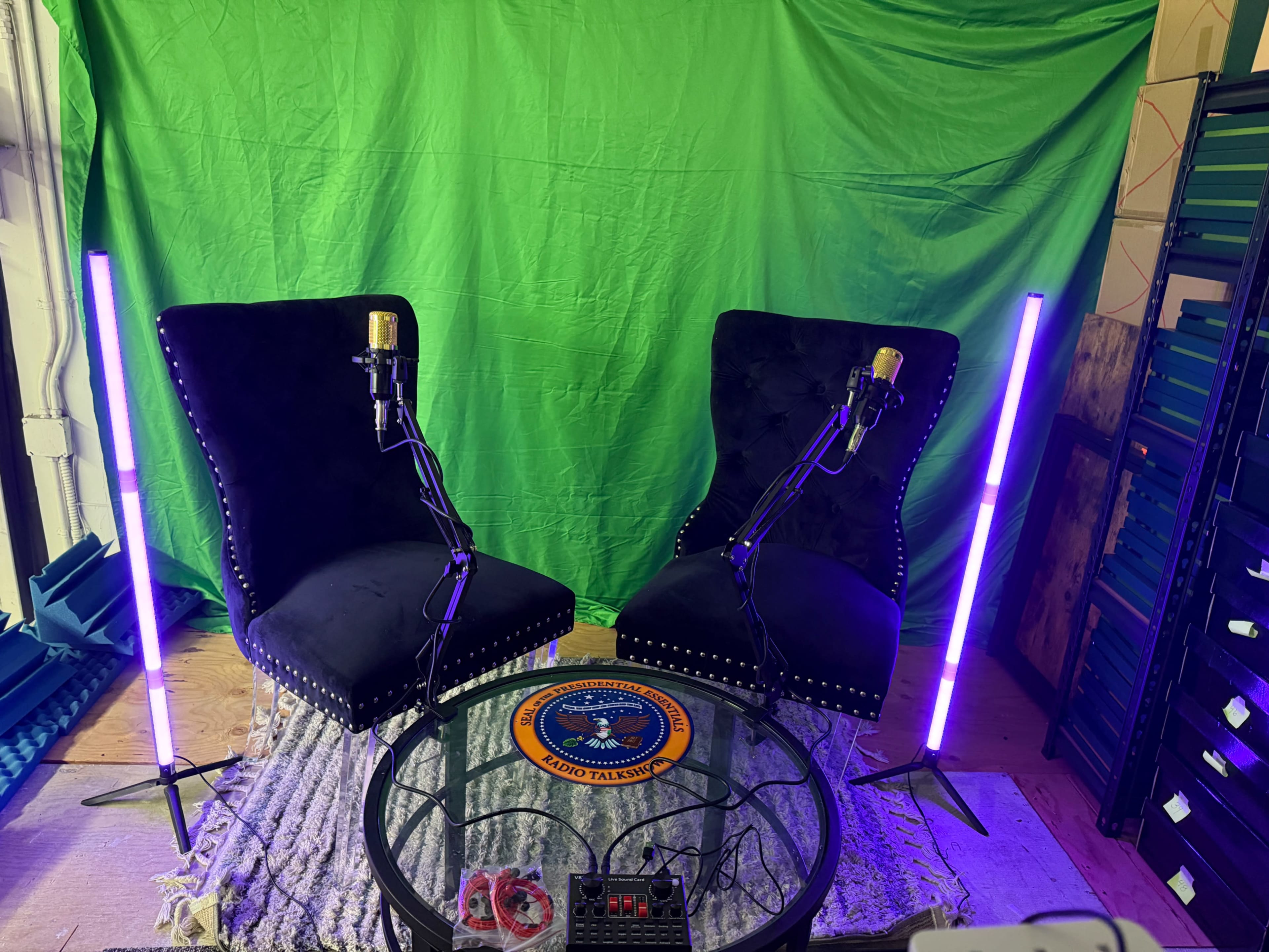 The scene features two black armchairs with microphones positioned on them, set against a green backdrop, with two illuminated purple light stands on either side of a round glass table.