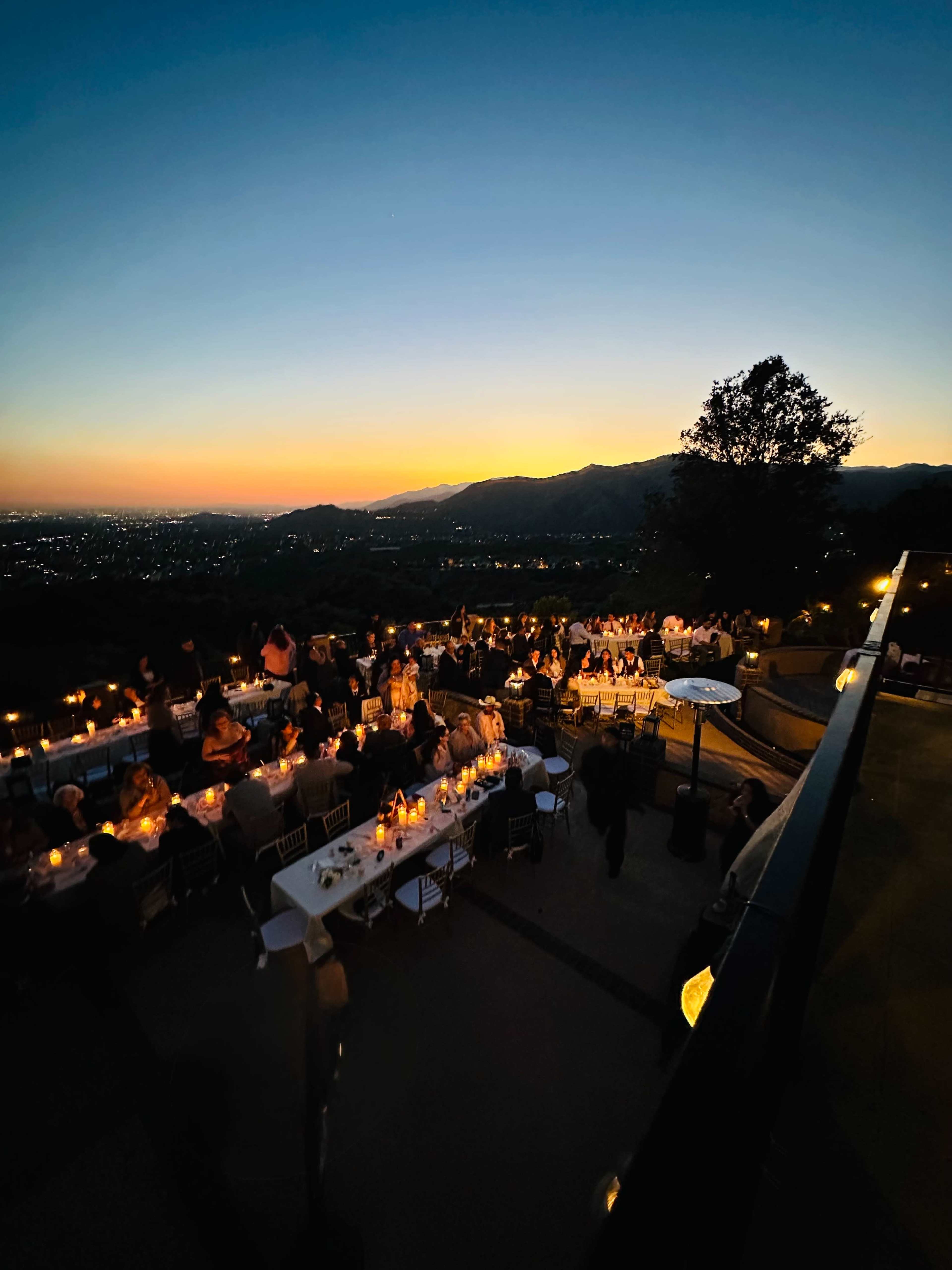A large outdoor dining event is taking place on a terrace with tables set for dinner, against a backdrop of a sunset over distant mountains.