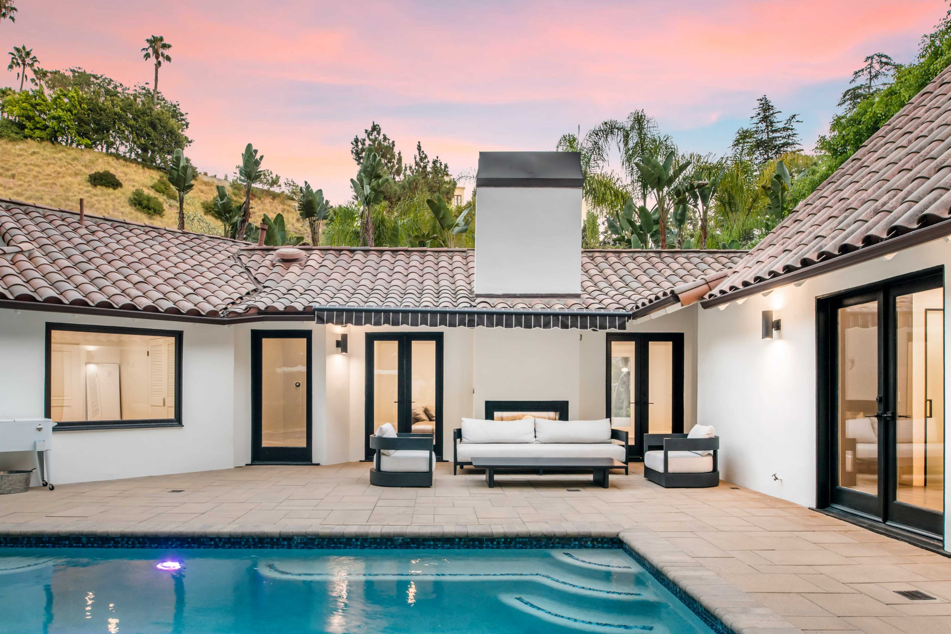 A modern outdoor space features a swimming pool surrounded by seating and a home with large windows and a tiled roof under a colorful sunset sky.
