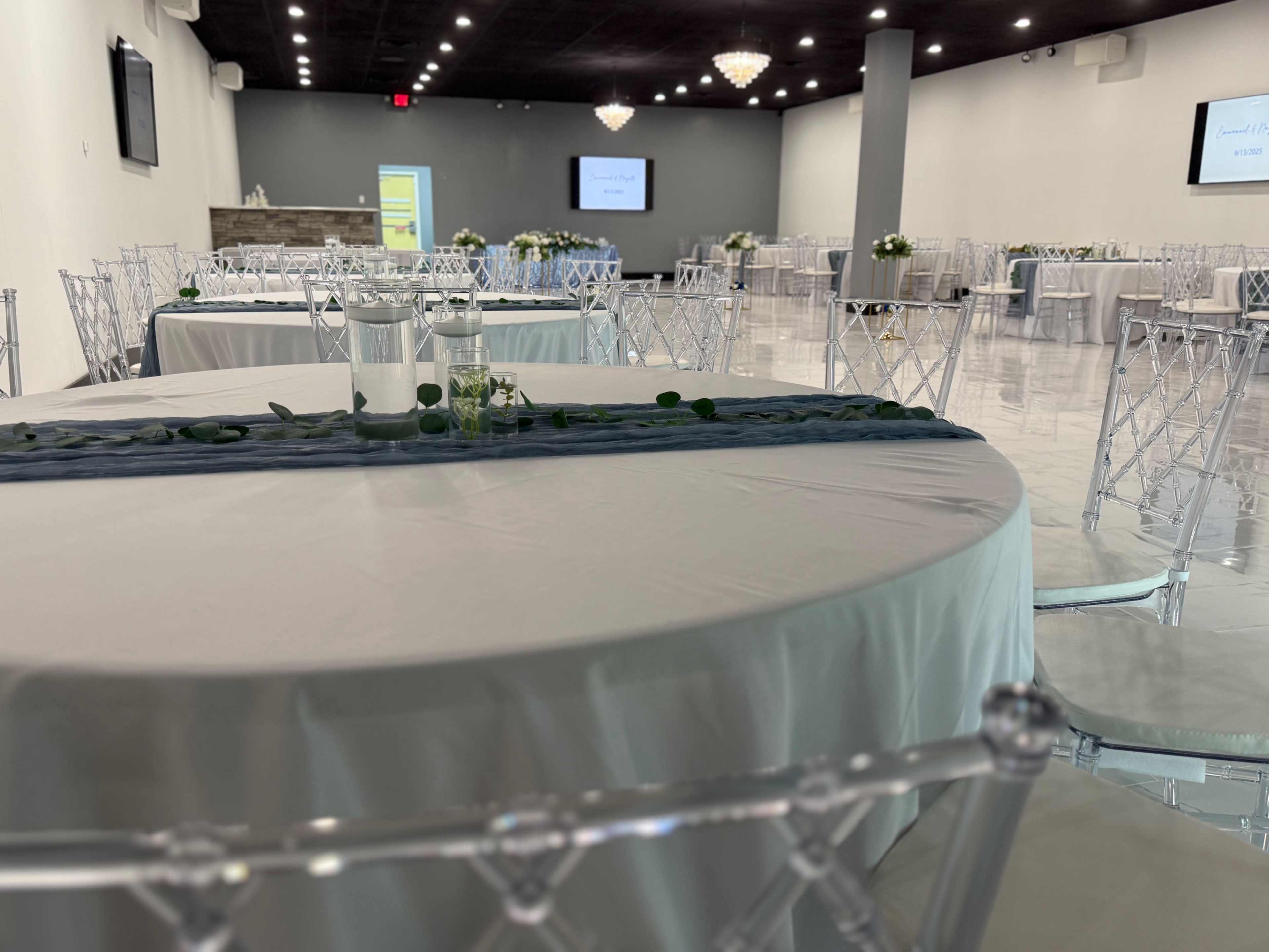 The image shows an upscale event space with multiple round tables set with light-colored tablecloths and transparent chairs, arranged for a gathering.