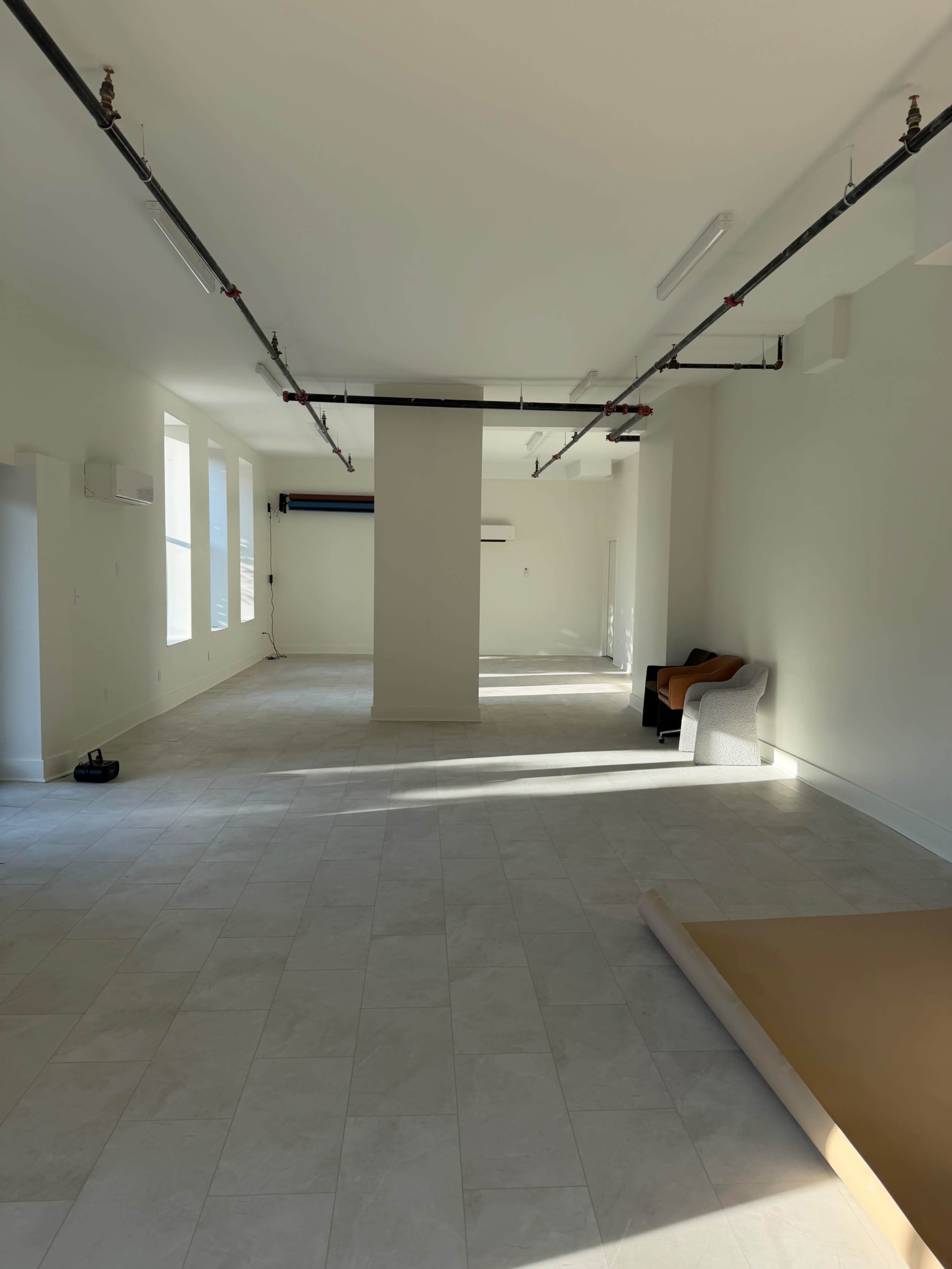 The image shows a spacious, empty interior room with white walls, large windows, and minimal furniture.