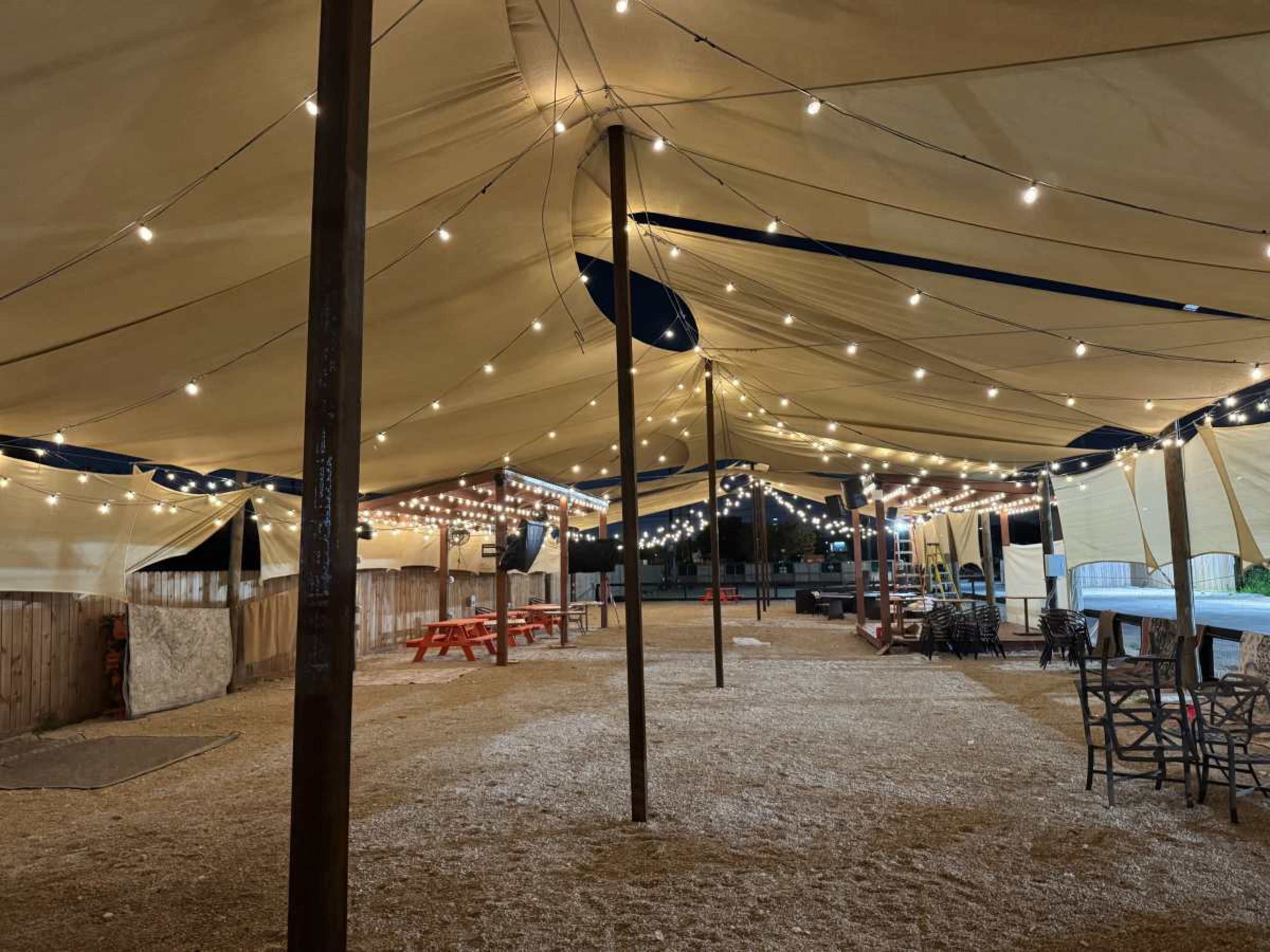 The image shows a large, open tent decorated with string lights, featuring wooden picnic tables and a gravel floor.