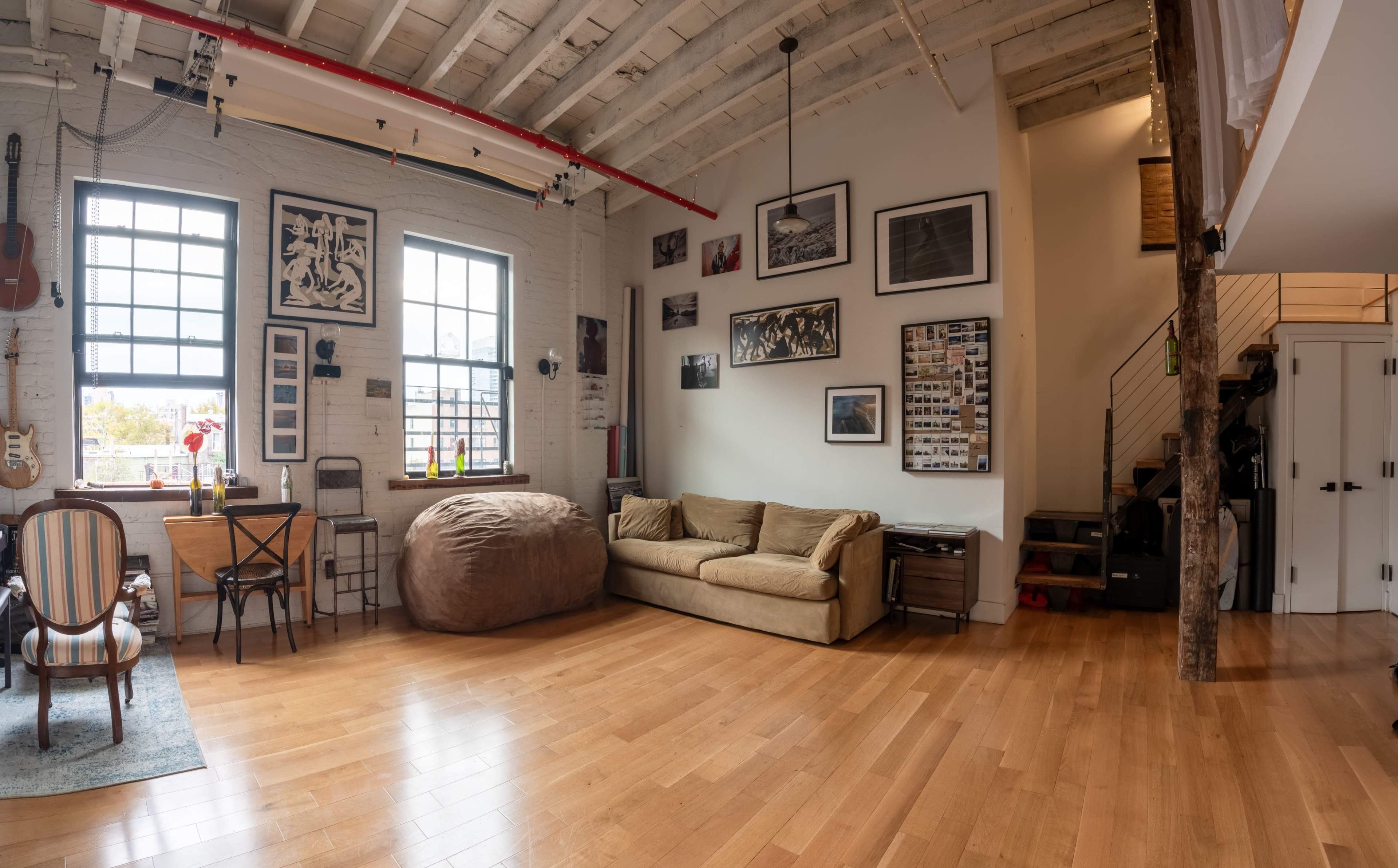 A spacious room features wooden floors, large windows, a sofa, a bean bag, a dining table, and walls adorned with various framed photos and artwork.