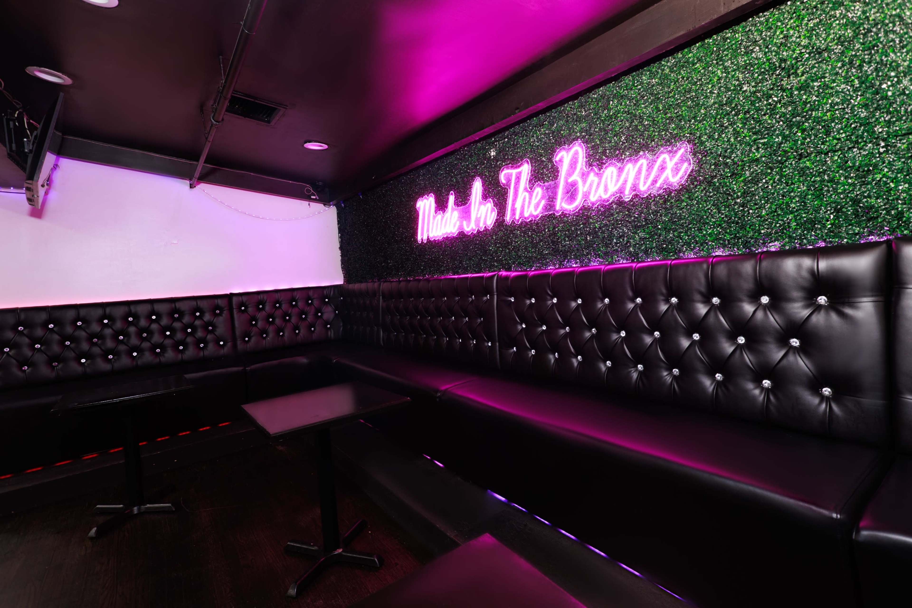 The image shows a small, dimly-lit lounge area with black tufted seating and a vibrant "Made in The Bronx" neon sign against a green wall.