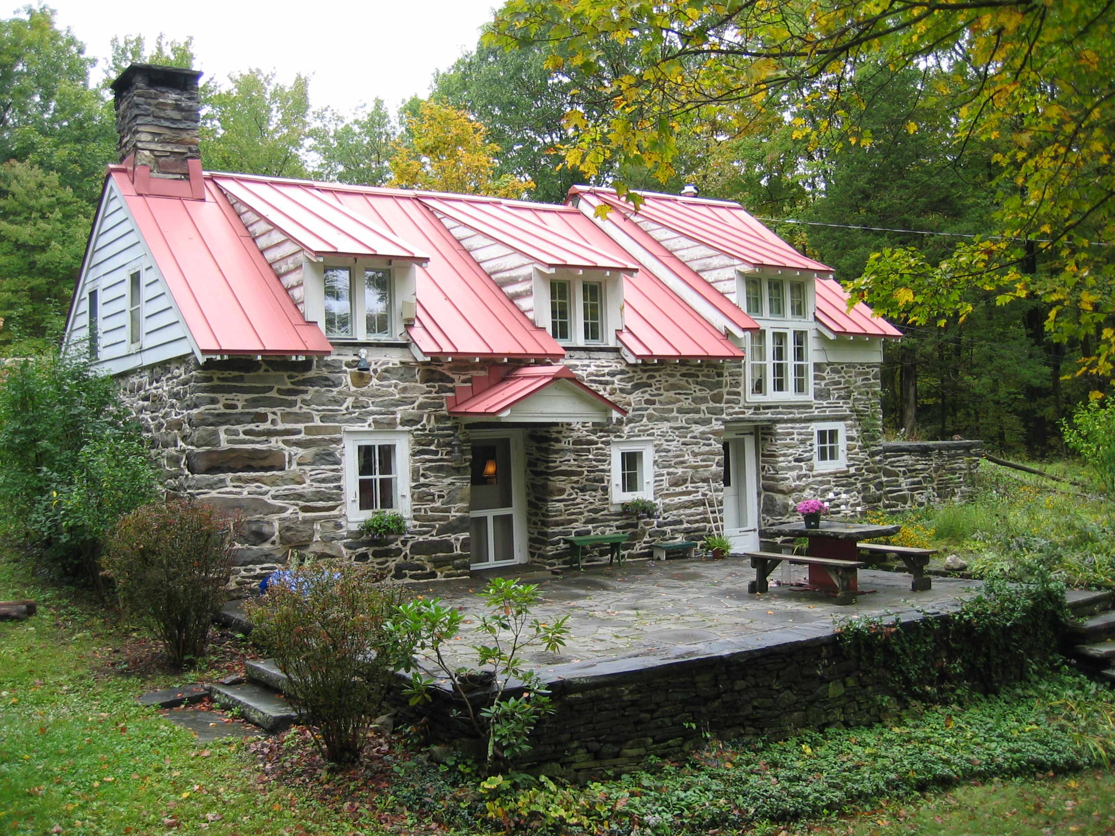 A stone house with a red metal roof and a small patio surrounded by greenery.
