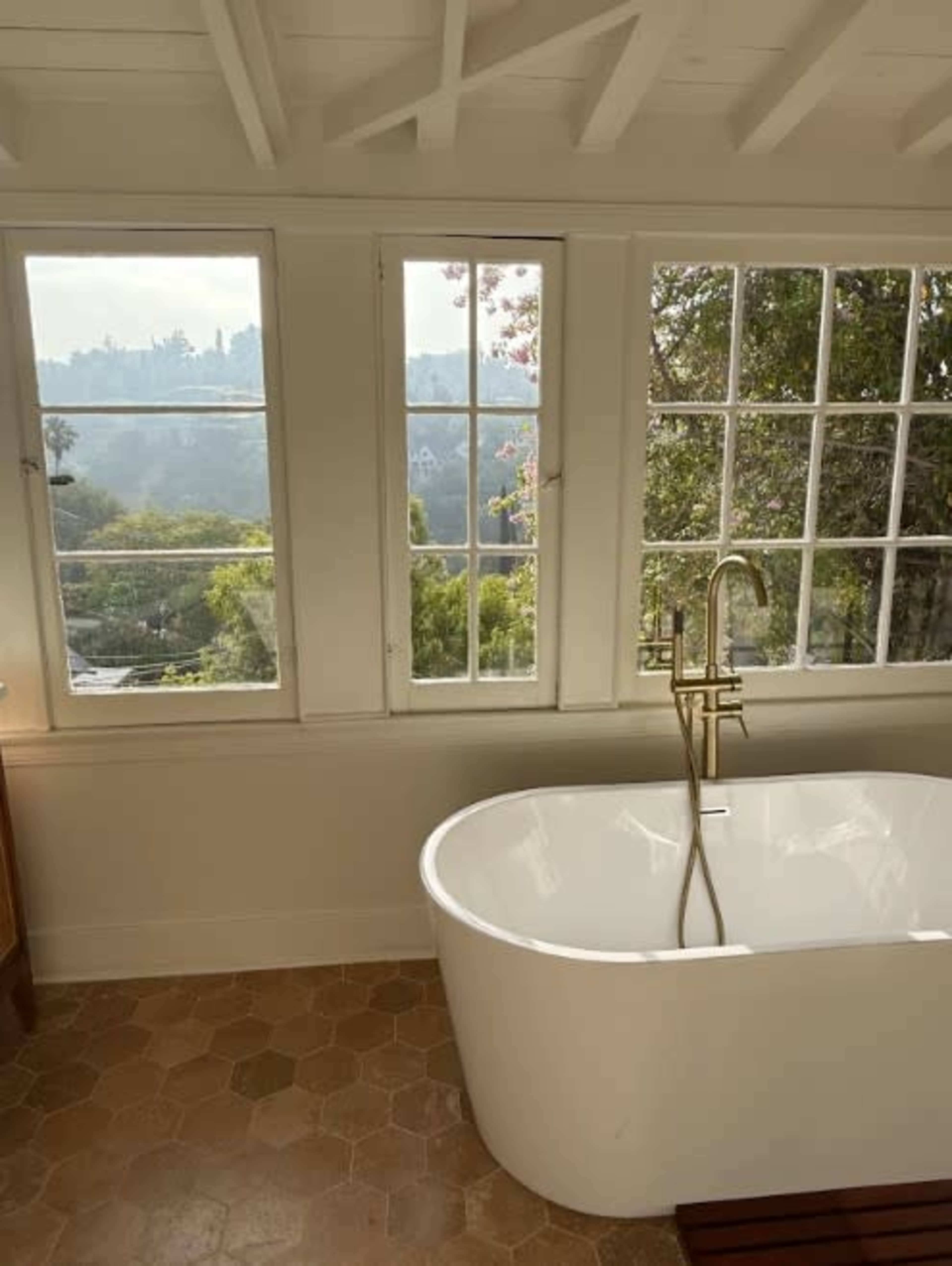 A modern bathtub is positioned beneath large windows that overlook a view of trees and hills.