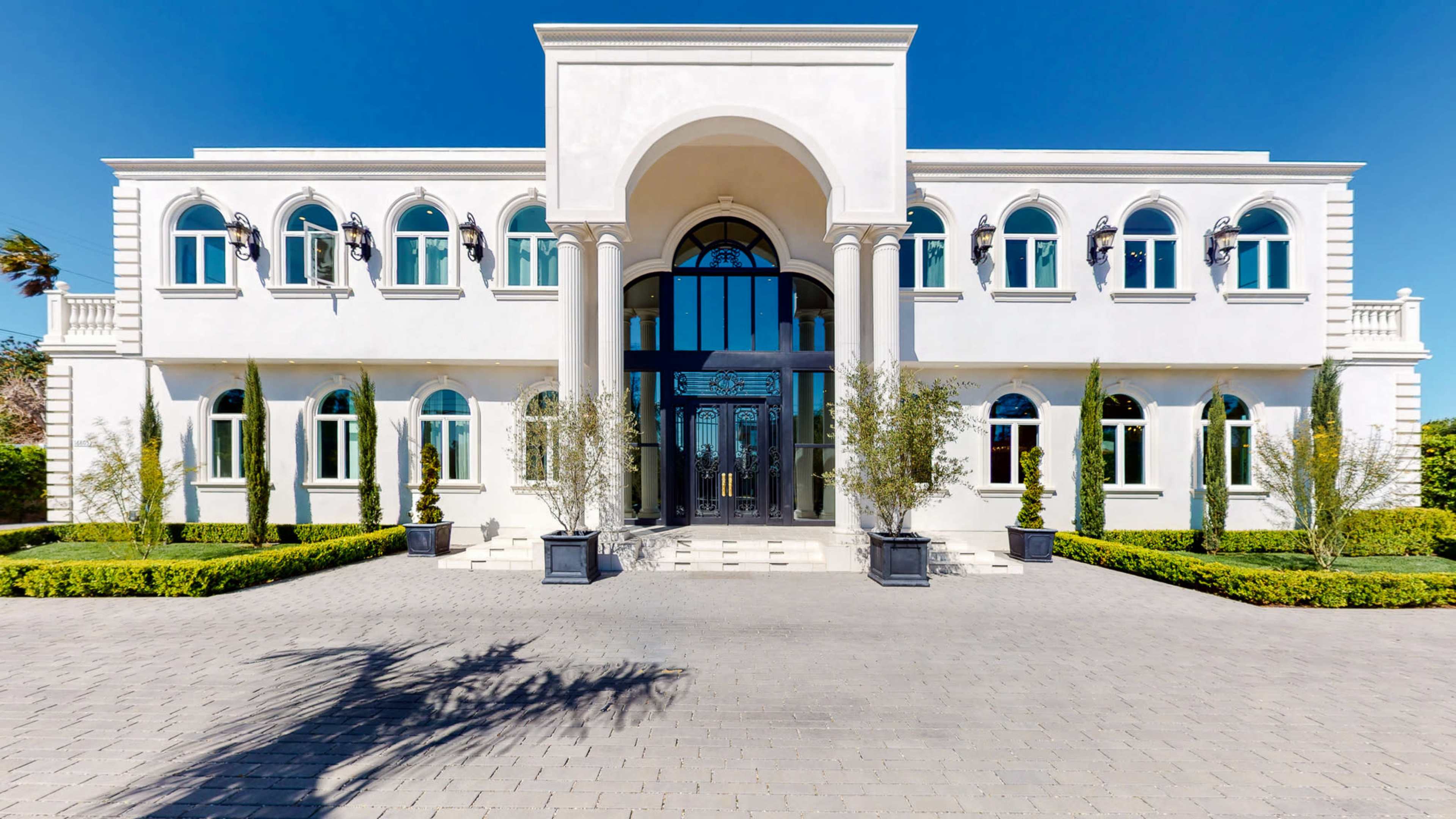 A large, modern mansion with a grand entrance, featuring arched doorways and symmetrical windows, is set among neatly landscaped greenery.