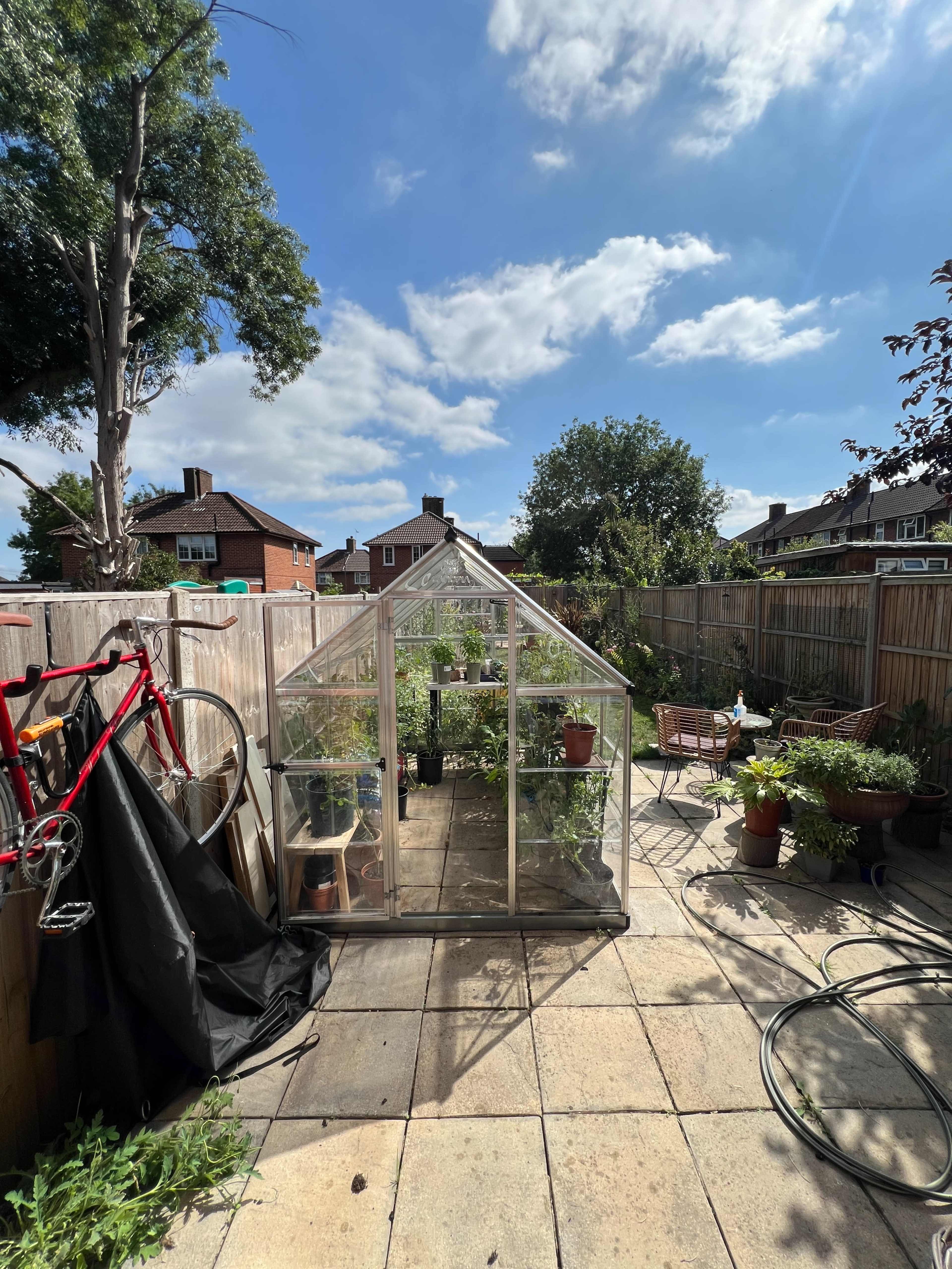 A greenhouse stands in a patio area surrounded by potted plants and outdoor furniture under a partly cloudy sky.