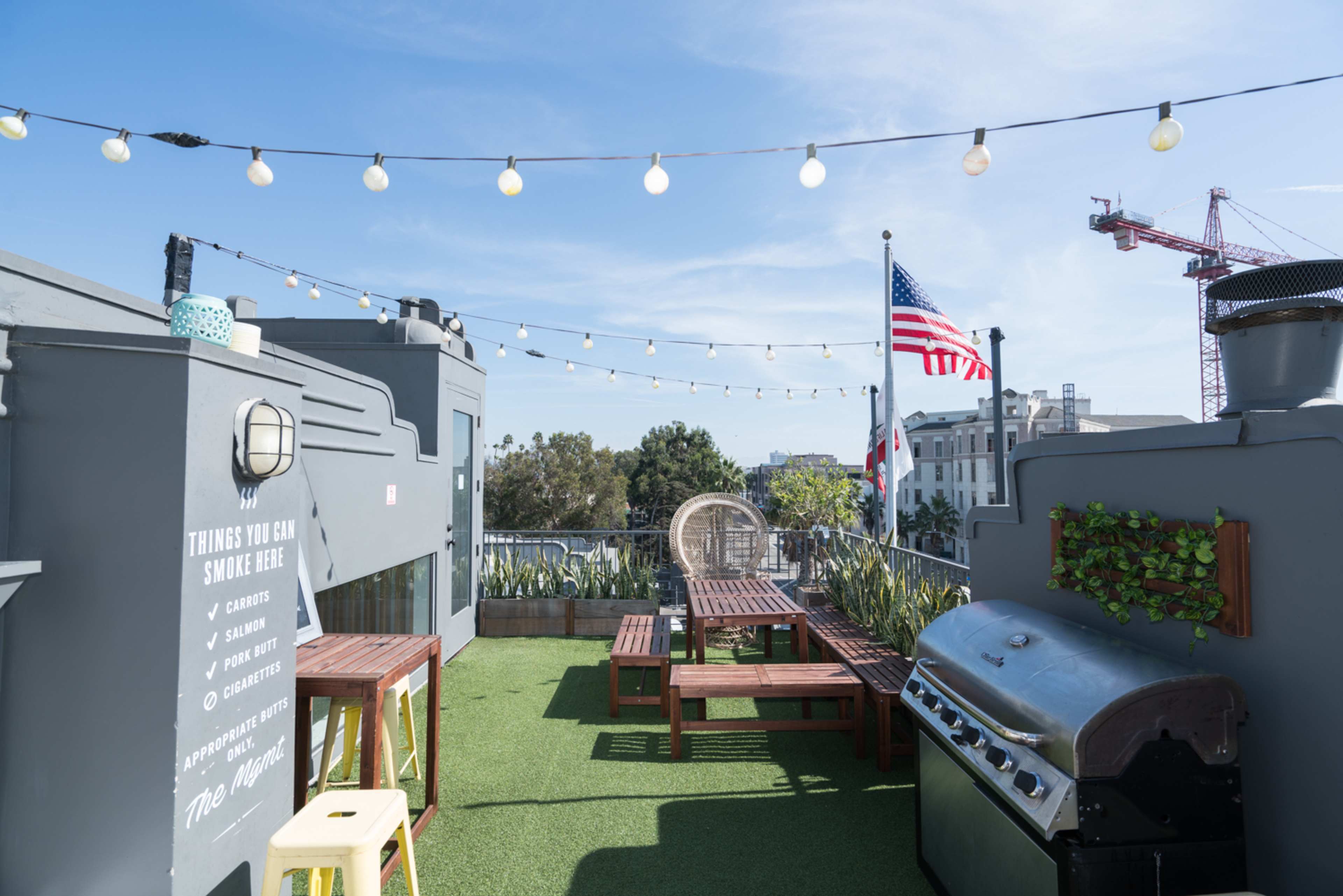 A rooftop area features string lights, seating arrangements, a barbecue grill, and a view of a flag and nearby buildings.