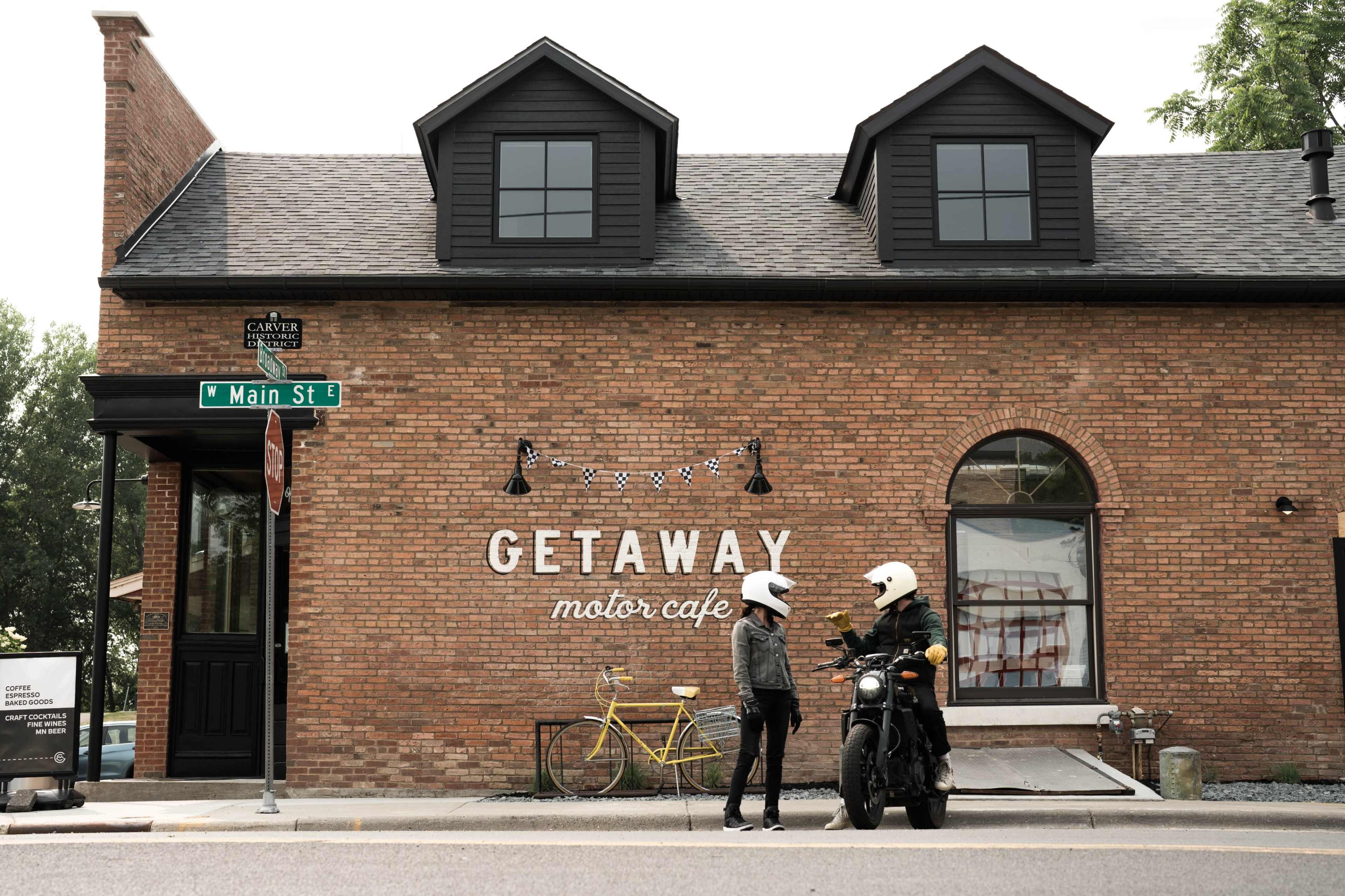 A woman in a helmet chats with a man in a motorcycle helmet outside the Getaway Motor Cafe, which features a brick building and decorative signage.