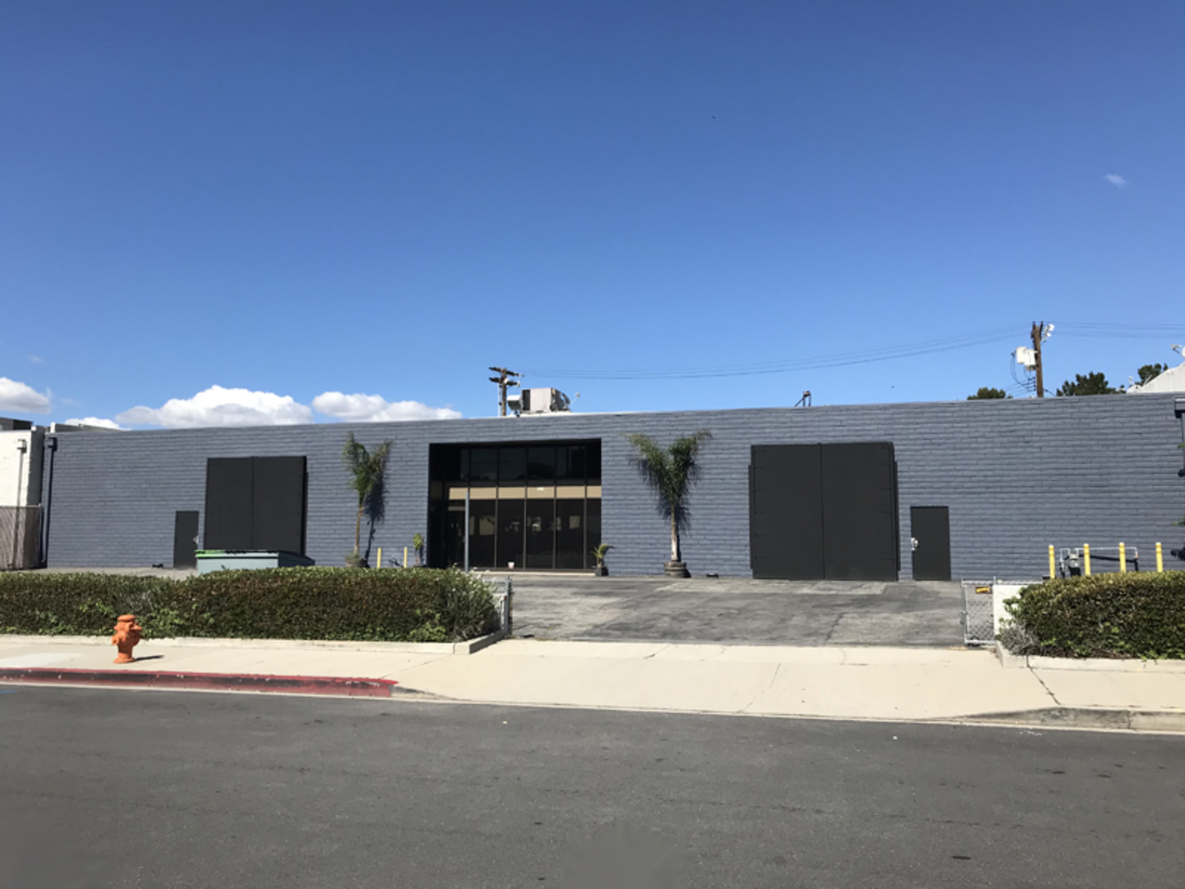 A gray commercial building with large black doors and palm trees flanks the entrance in a clear blue sky.
