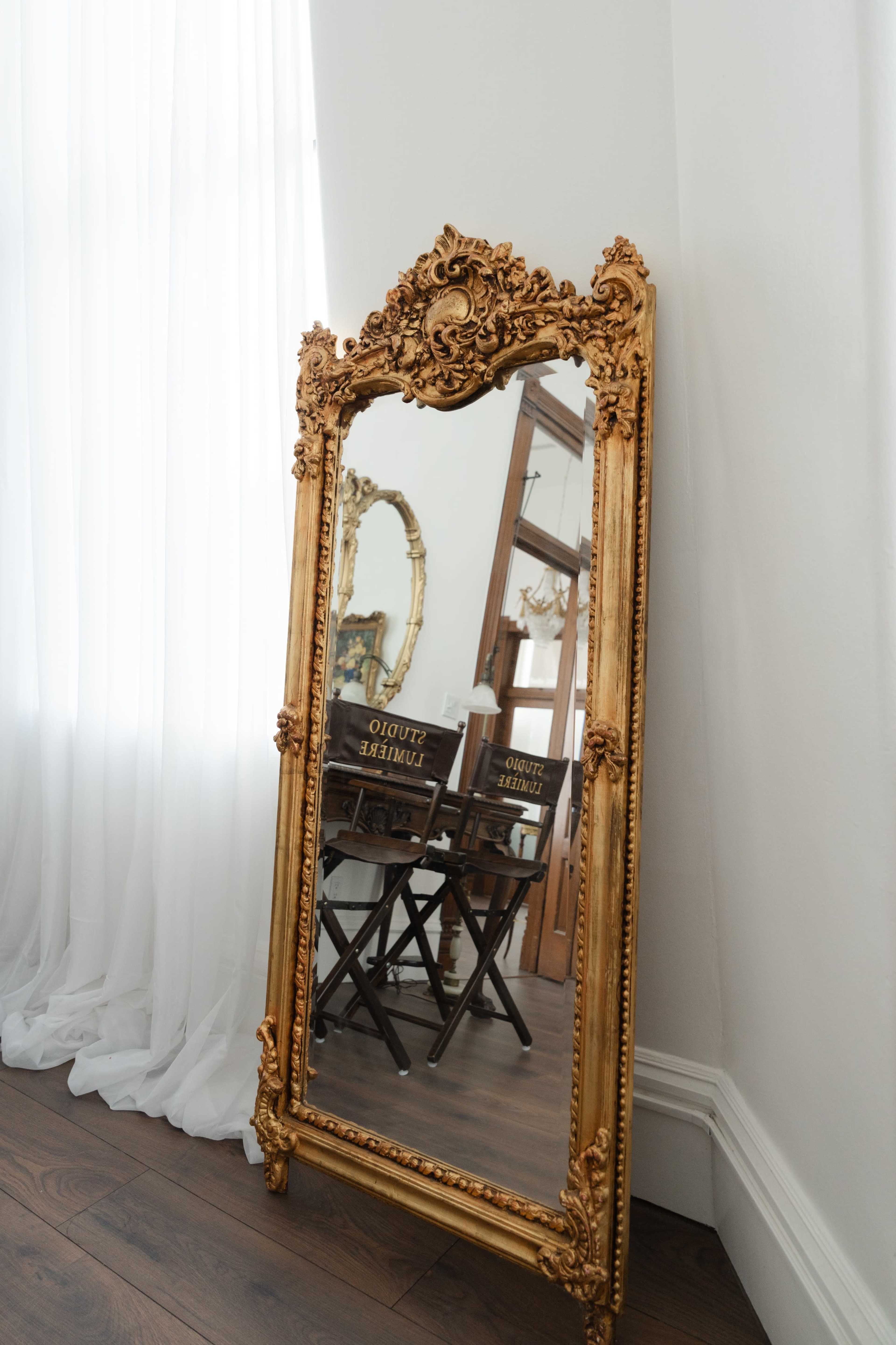 A large ornate gold-framed mirror leans against a wall, reflecting a wooden chair and part of a curtain.