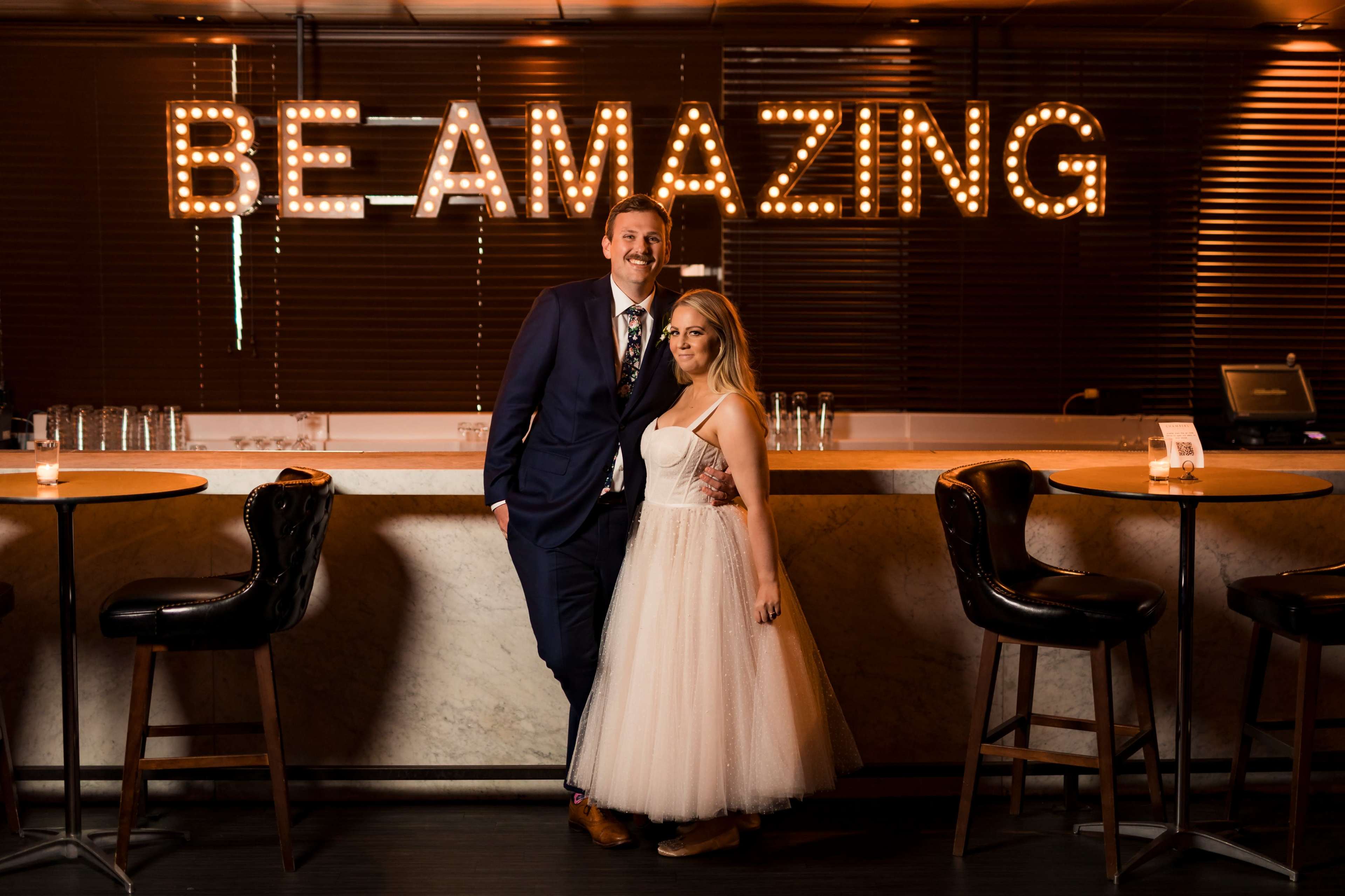 A man in a suit and a woman in a white dress stand together in front of a bar with illuminated letters spelling "BE AMAZING."