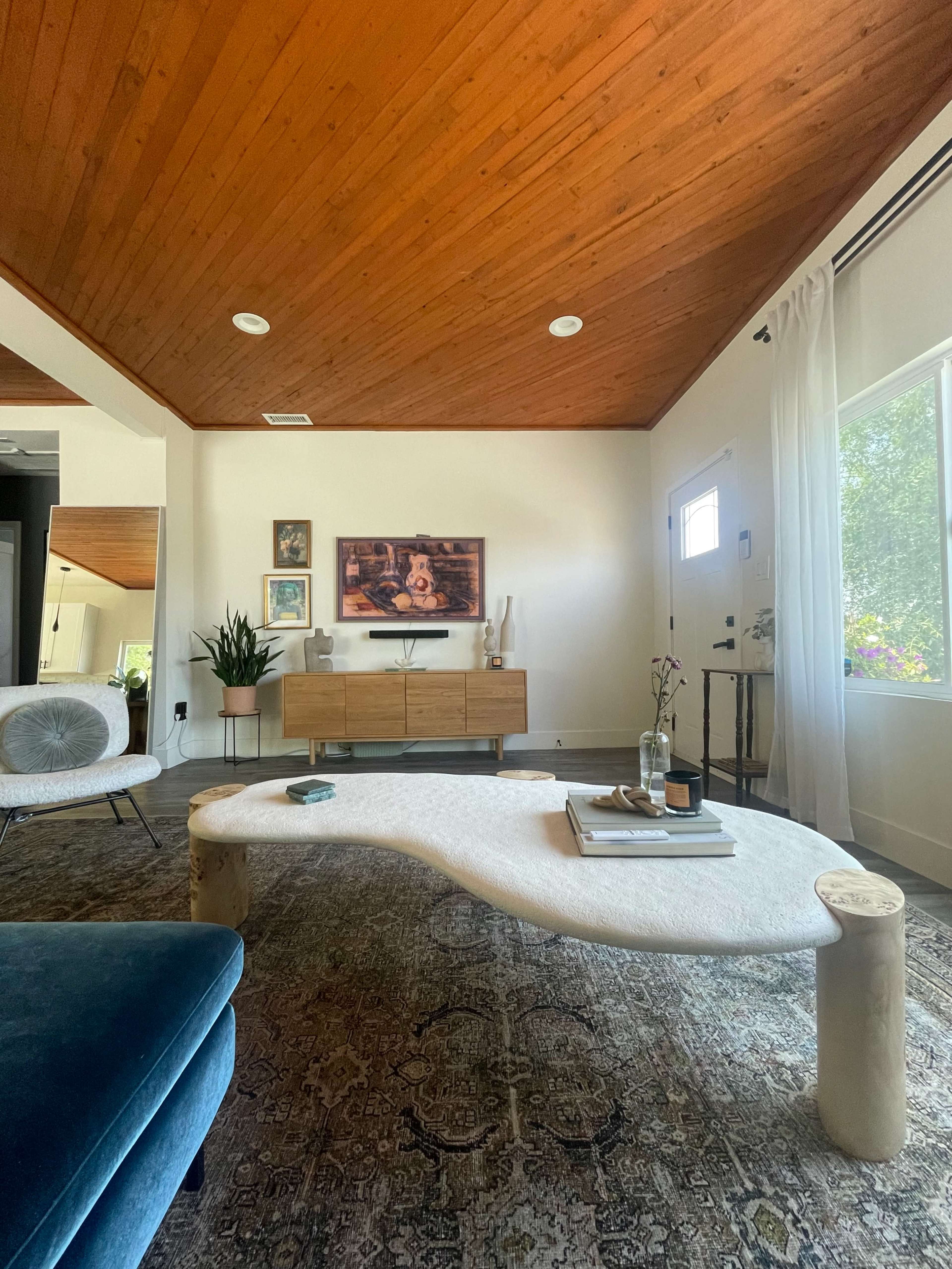The living room features a wooden ceiling, a large area rug, a low coffee table, a media console with a television, and a door leading to the outside.