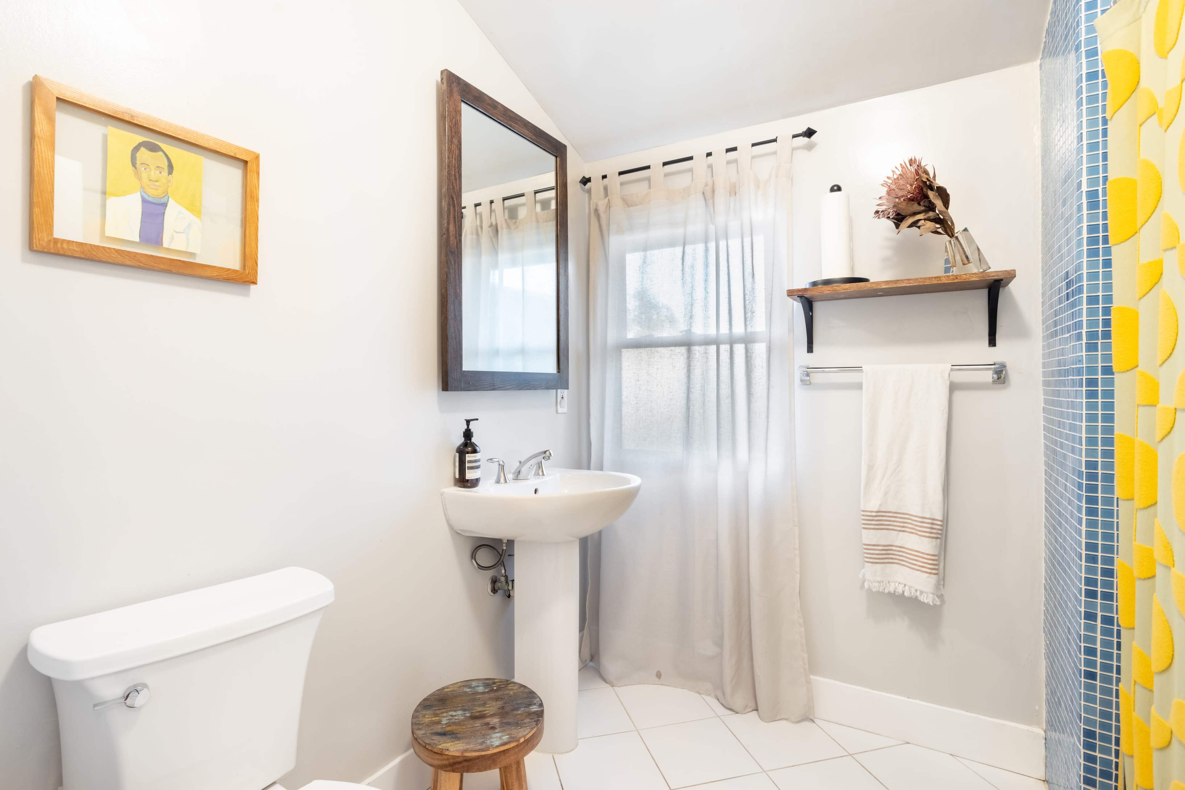 A small bathroom featuring a sink with a mirror, a toilet, a wooden stool, and a shower curtain with a blue tile pattern.
