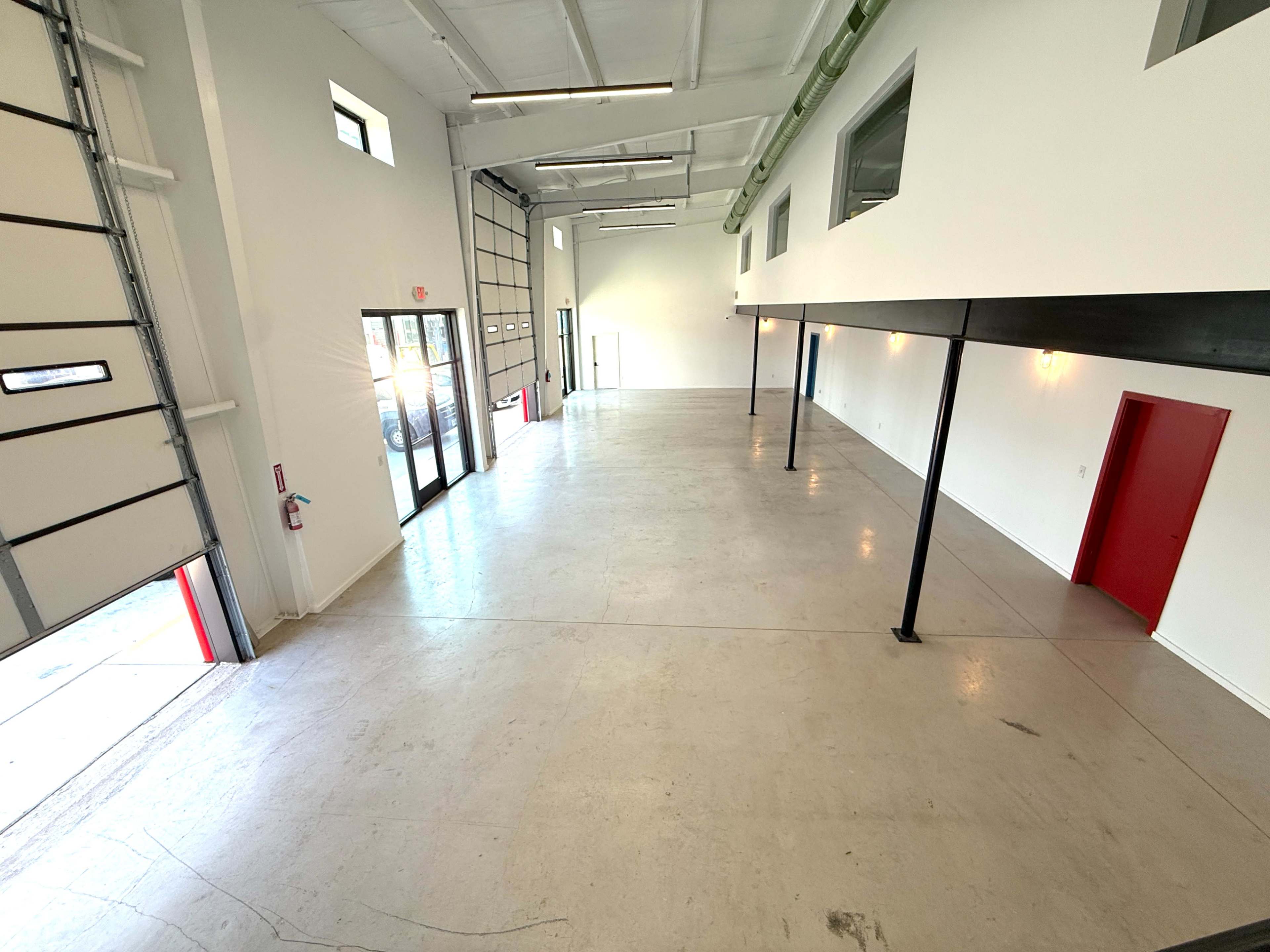 The image shows a large, empty commercial space with high ceilings, concrete floors, and red-accented doors.