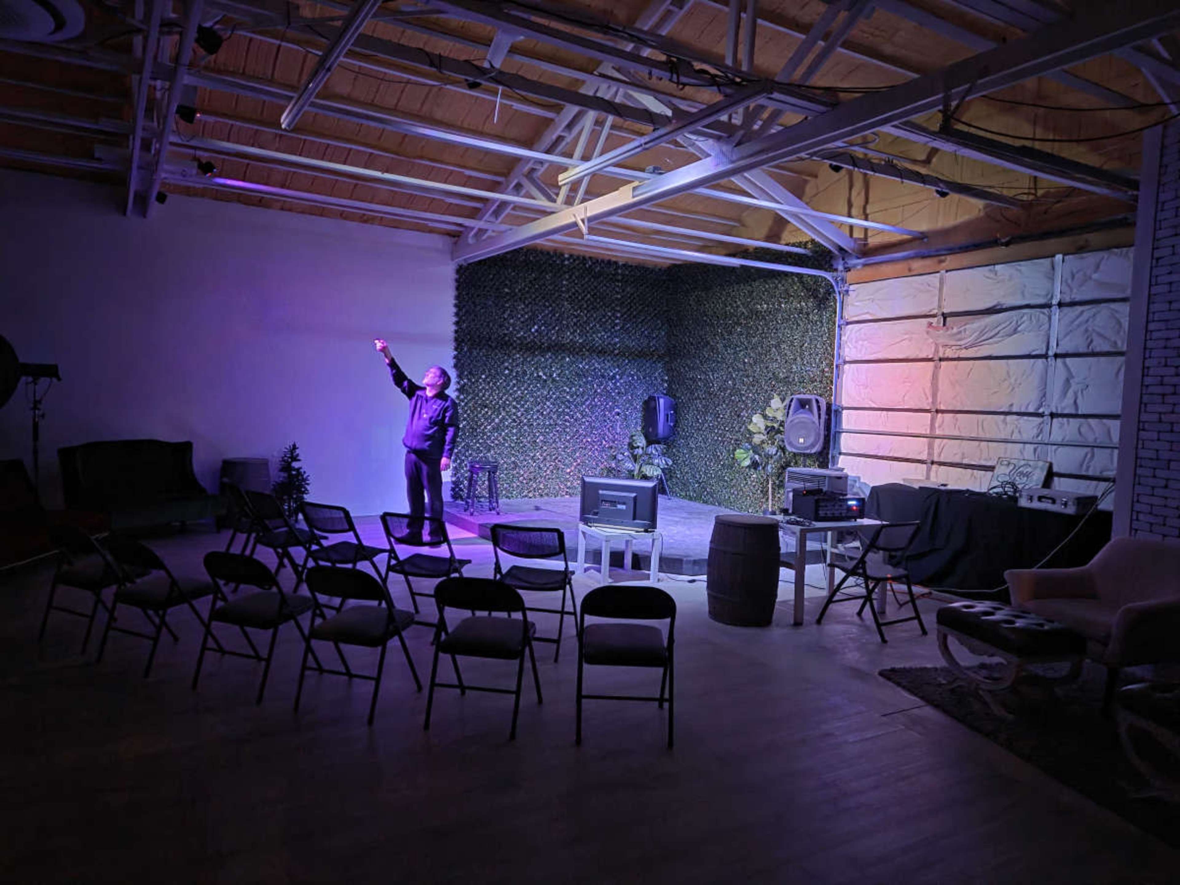 A person stands on a small stage in a sparsely furnished performance space with rows of chairs facing the stage and colorful lights illuminating the backdrop.