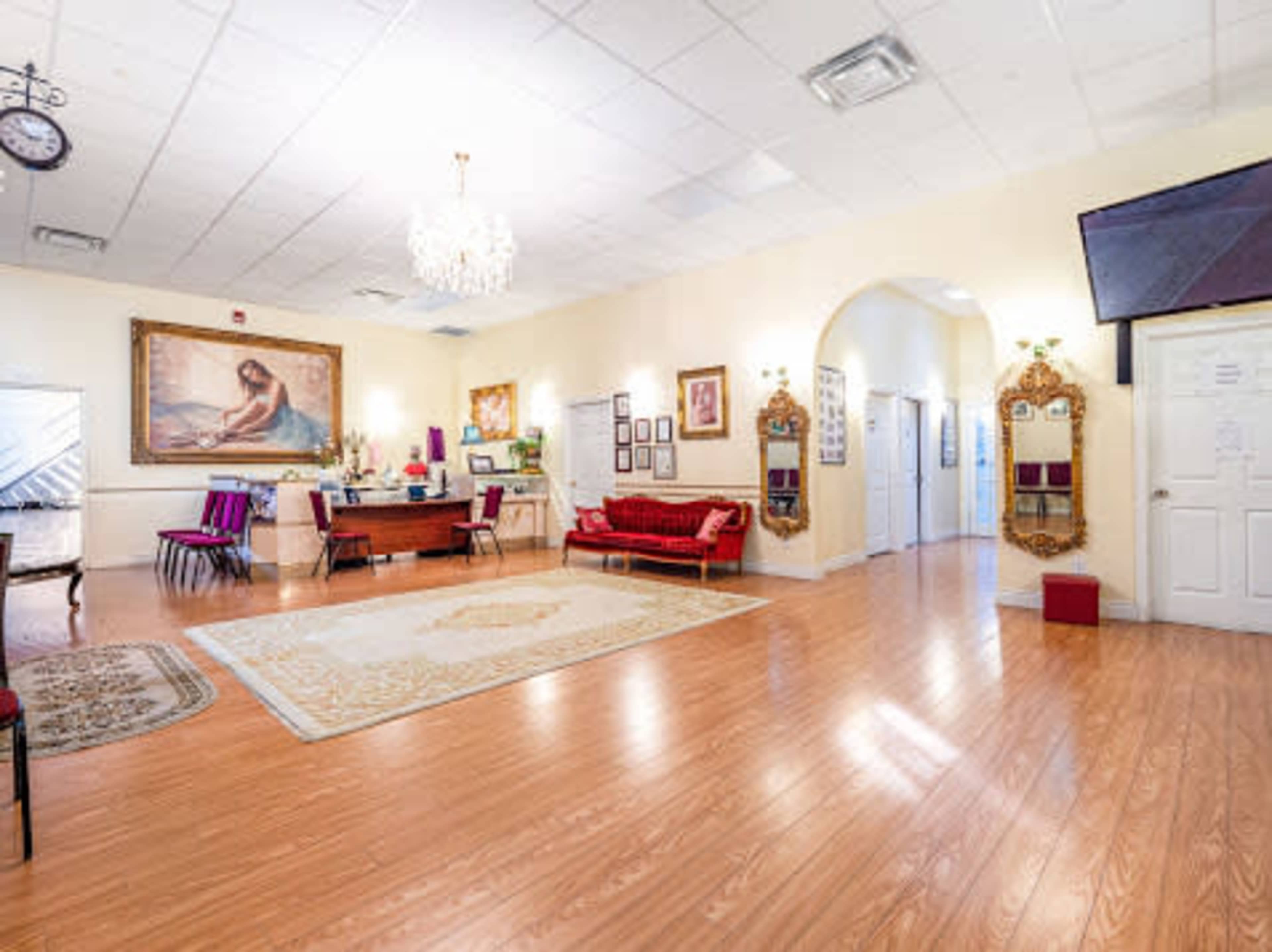 A spacious room with wooden flooring, featuring a chandelier, a large painting, various seating areas, and multiple doorways leading to adjacent rooms.