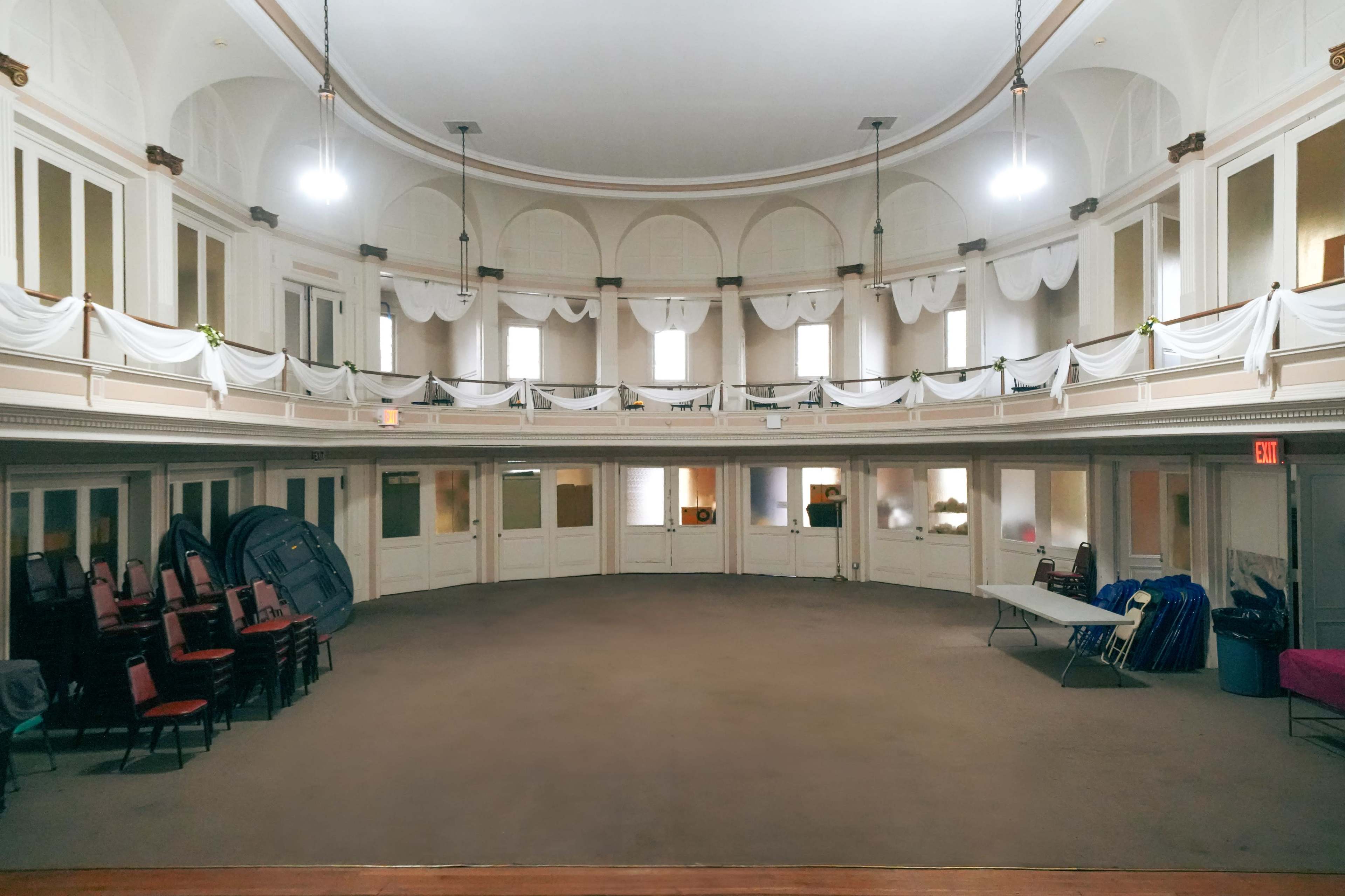 A spacious, empty auditorium with a circular layout, featuring rows of stacked chairs on one side and white drapes along the upper walls.