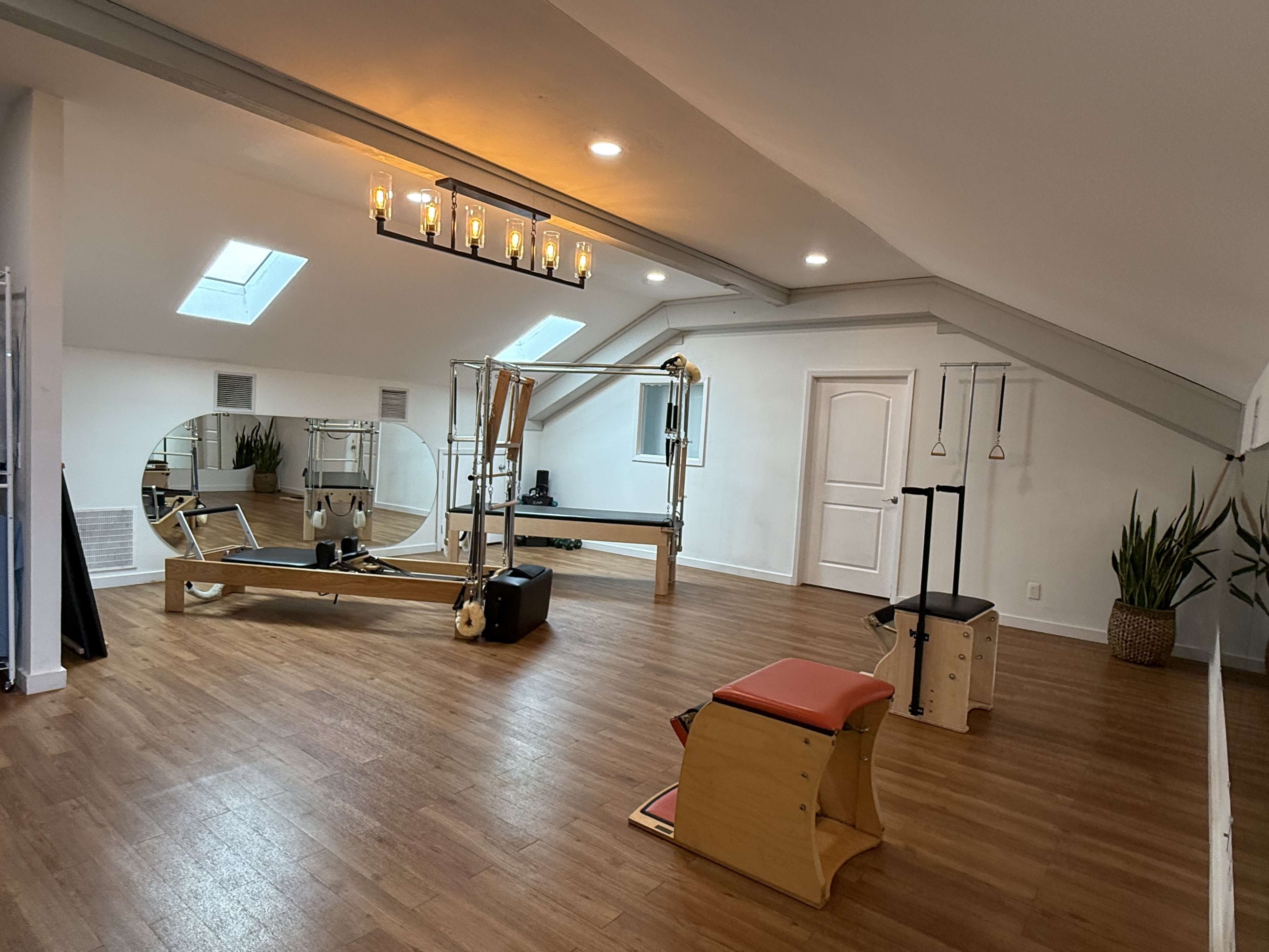 The image shows a spacious Pilates studio featuring a reformer, a chair, and various exercise equipment arranged on wooden flooring with a skylight and a large mirror.