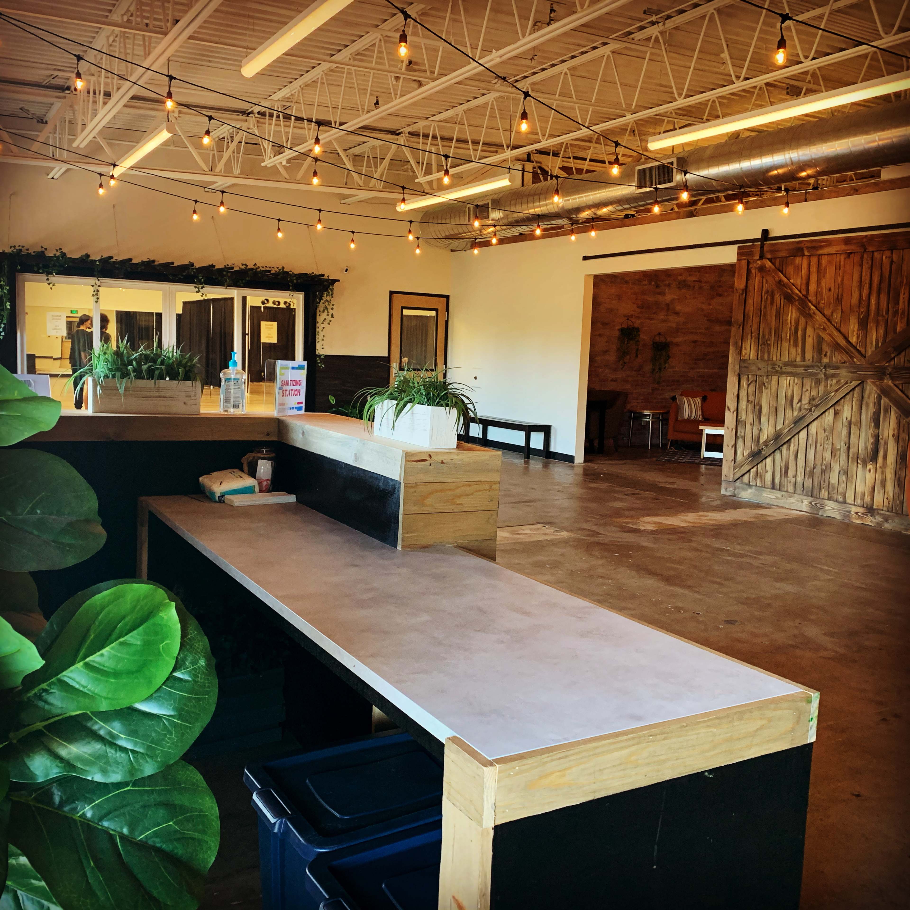 A spacious indoor area with a wooden counter, potted plants, exposed lighting, and a large wooden door leading to another room.