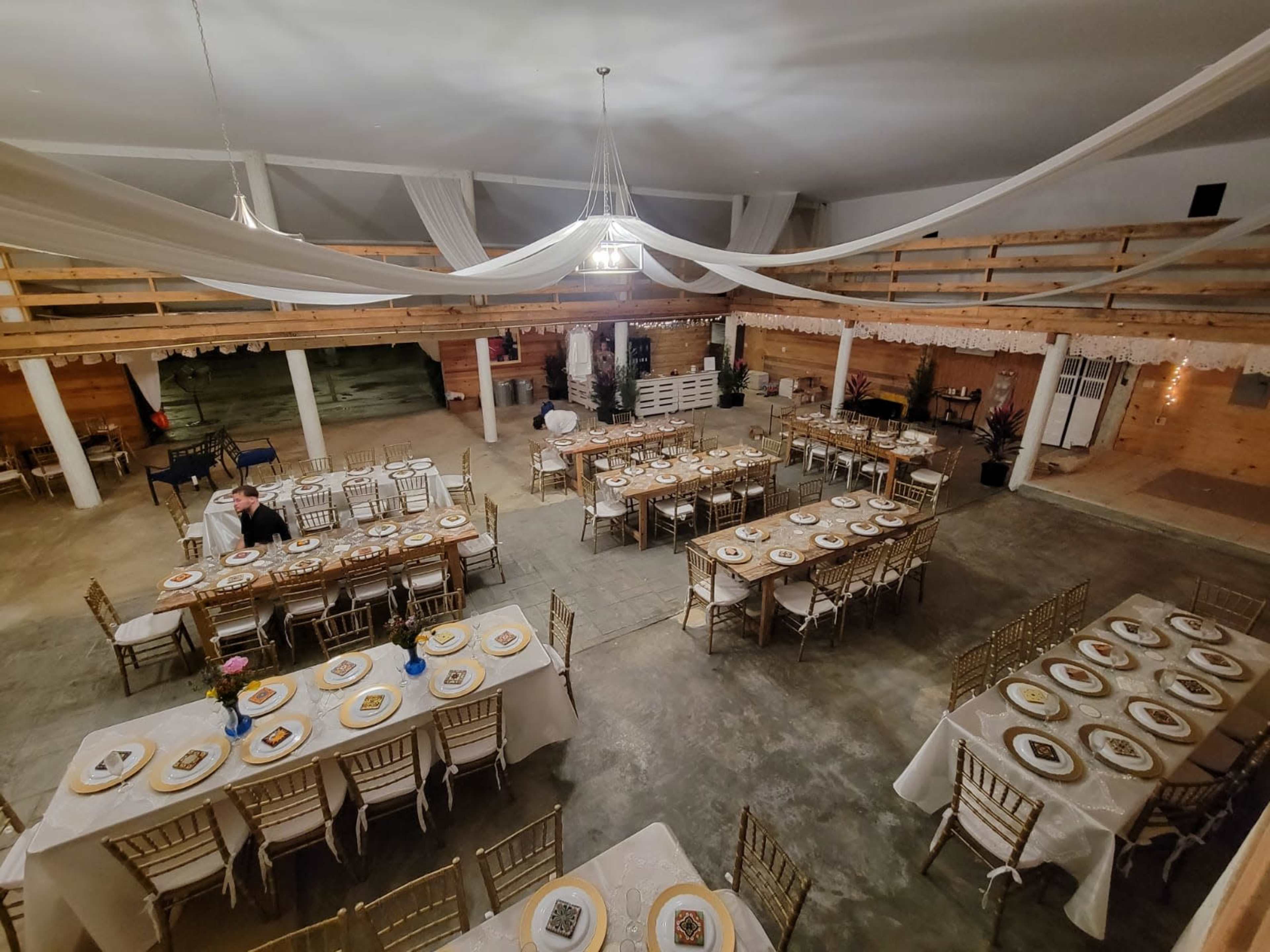 The image shows a spacious event hall set with multiple tables arranged for a dining event, featuring gold-colored chairs and table settings.