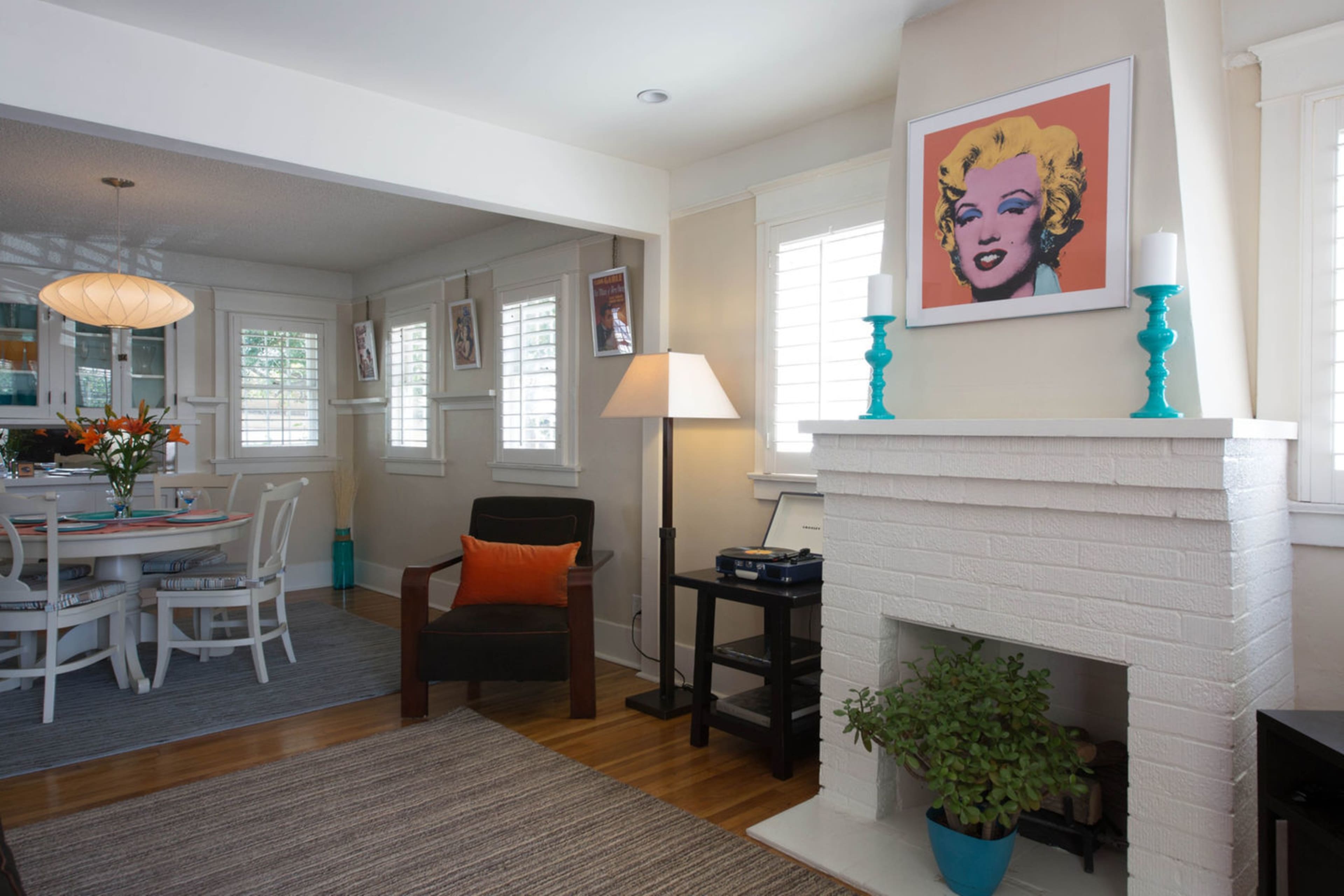 The image depicts a well-lit living room with a white-brick fireplace, a chair with an orange cushion, and a dining area visible in the background.