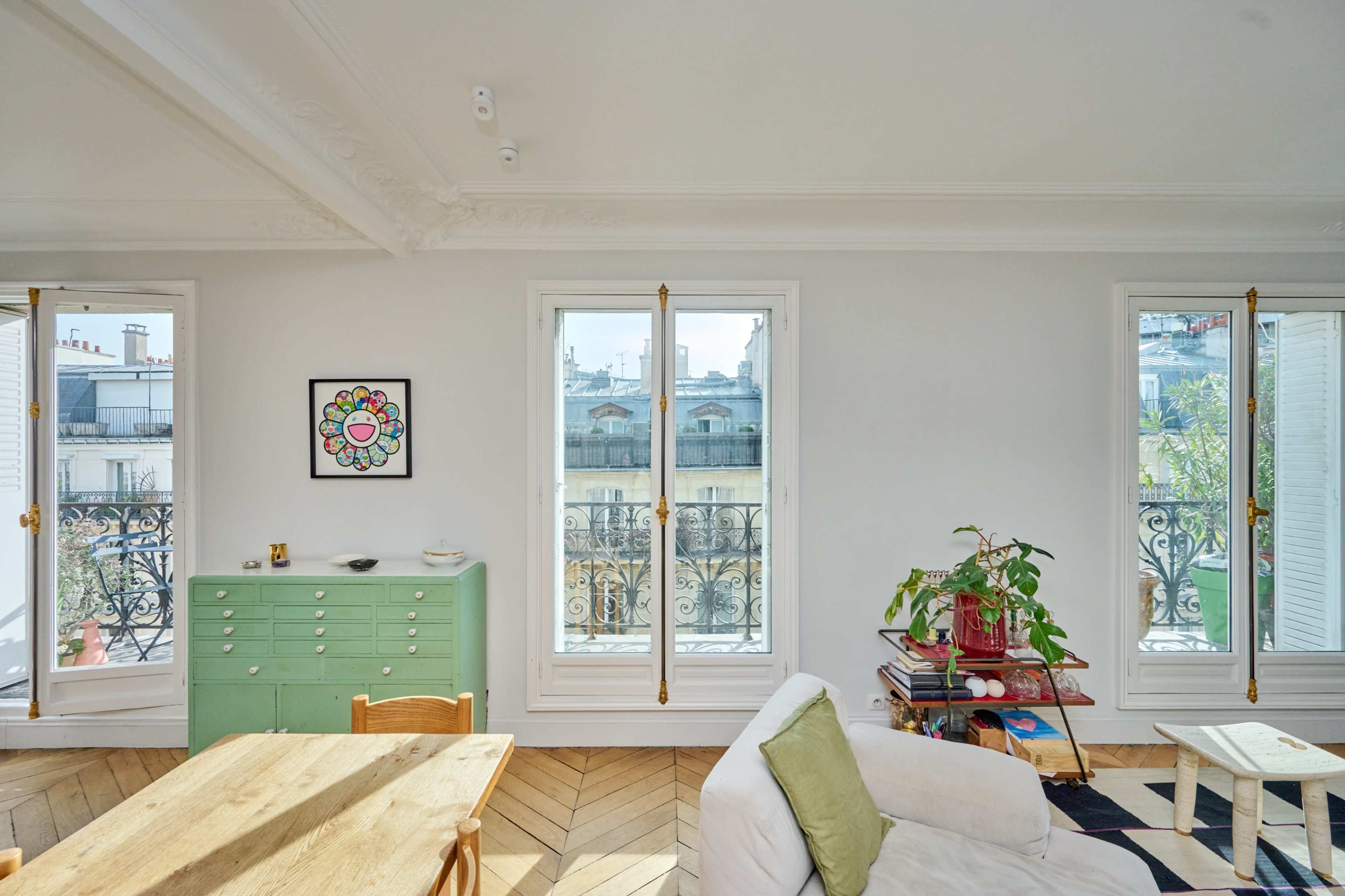 A bright living room features a green dresser, a wooden dining table, and large windows with decorative railings overlooking an outdoor view.
