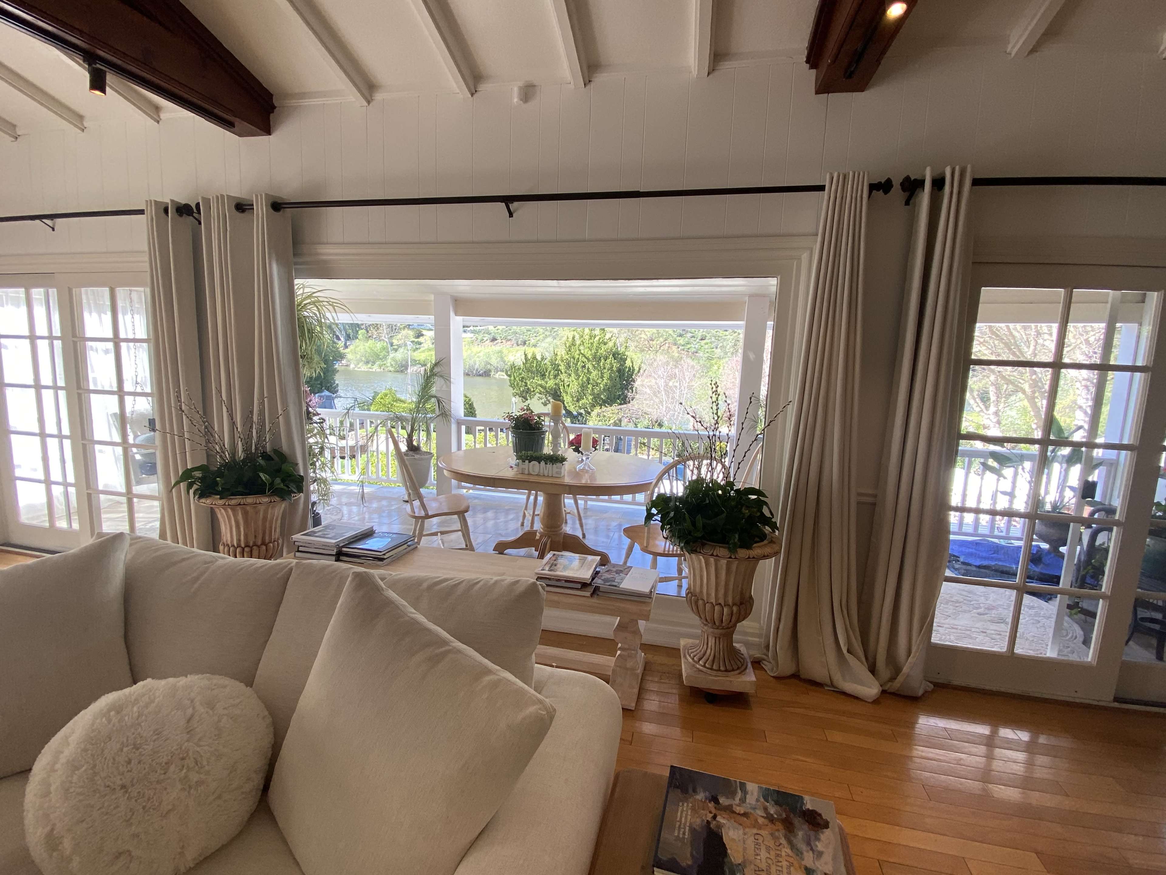 A cozy living room features a large white sofa and opens through sliding glass doors to a terrace with a dining table and lush greenery beyond.