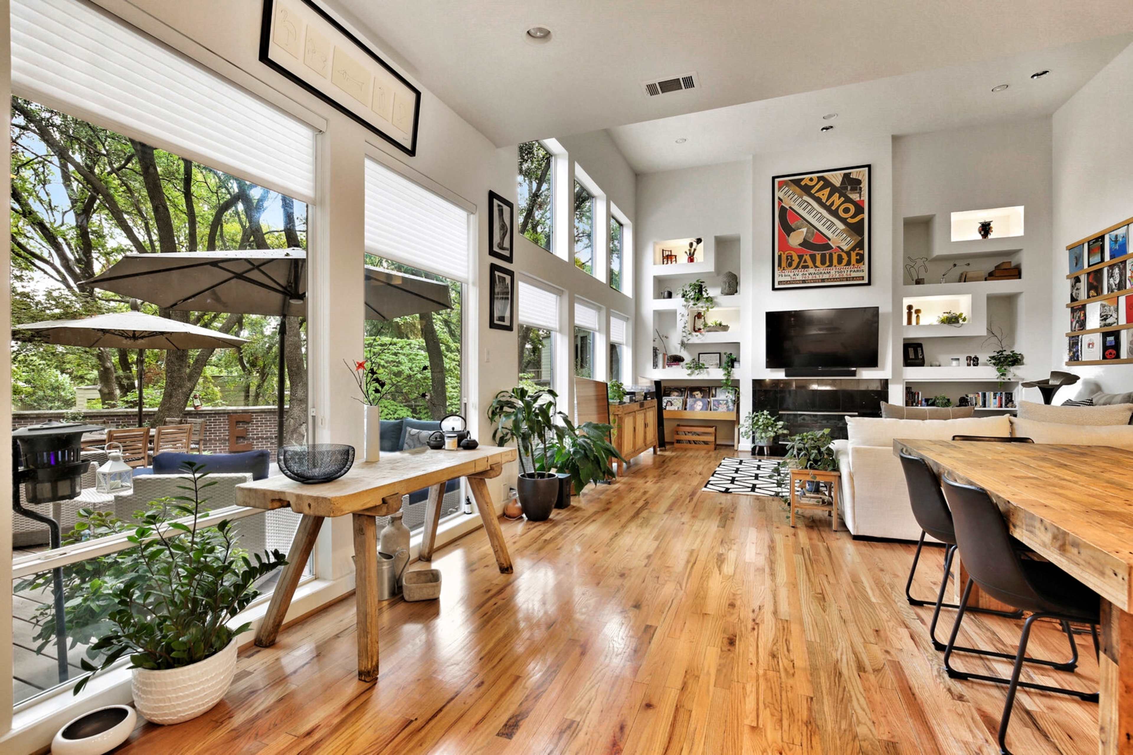 The room features large windows, wooden flooring, and a mix of furniture, including a dining table and a seating area with plants and decorative items.